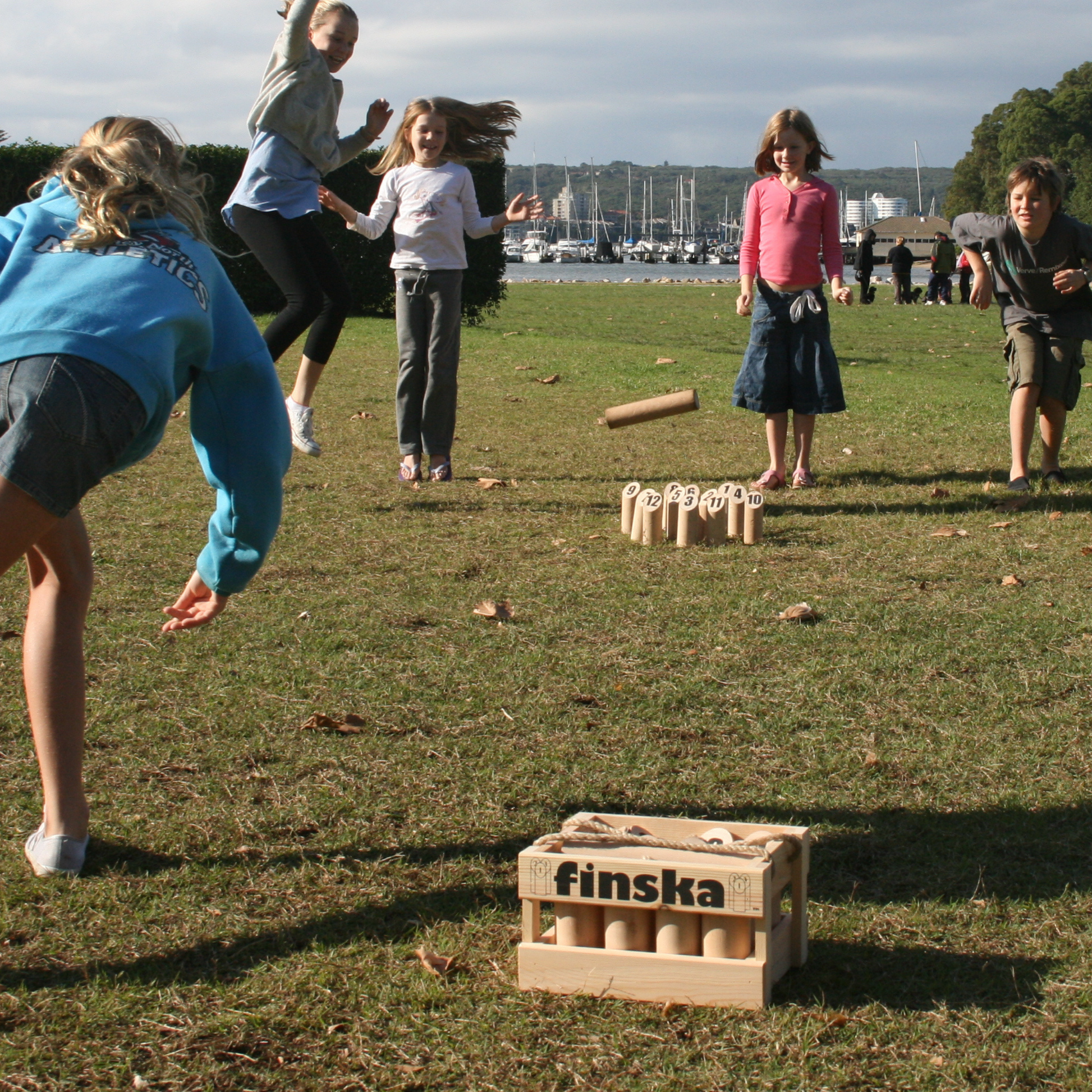 Shown kids playing on the grass the Original Finska natural birch outdoor throwing game set, includes 12 numbered pins, a Finska throwing log, birch carry crate, rule card, and score sheet. Traditional Finnish lawn game promoting strategy, skill, and fun for all ages.