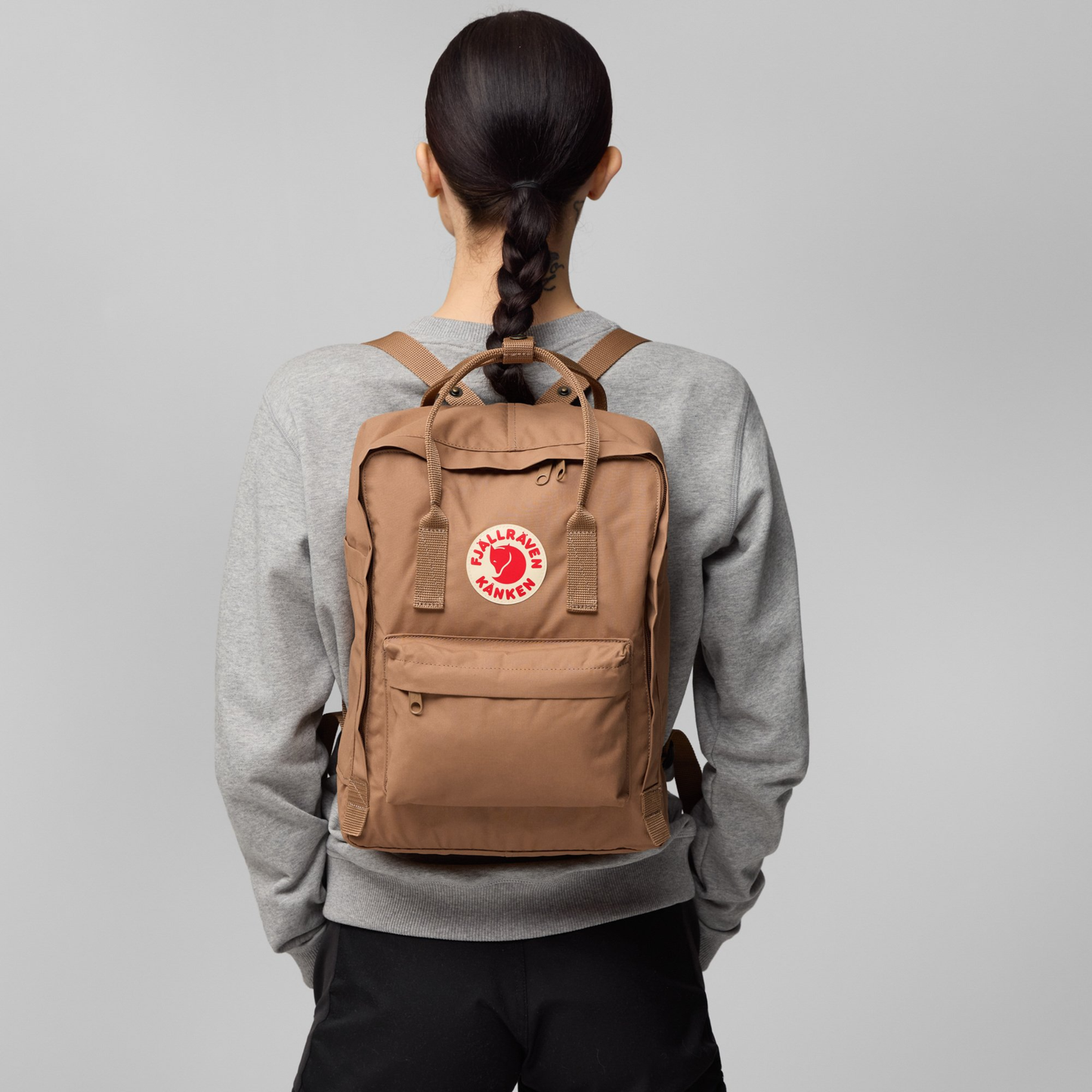Back view of woman wearing The Fjallraven Kanken Backpack which is made from hard-wearing Vinylon F fabric and features a spacious main compartment with a full-length zip, removable PE foam seat cushion, two flat side pockets, a zipped front pocket, adjustable shoulder straps, and top handles. Durable and practical backpack for everyday use.