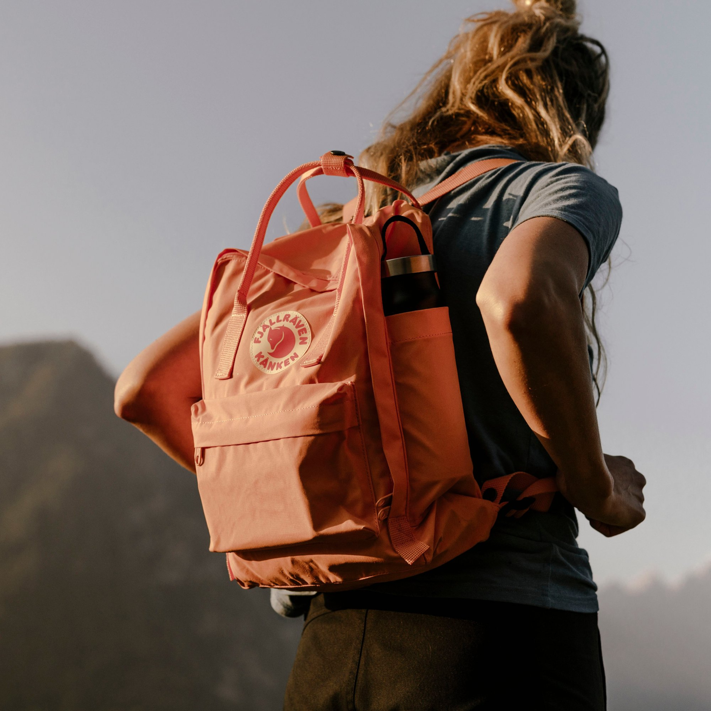 Showing a woman from the mountain wearing The Fjallraven Kanken Backpack which is made from hard-wearing Vinylon F fabric and features a spacious main compartment with a full-length zip, removable PE foam seat cushion, two flat side pockets, a zipped front pocket, adjustable shoulder straps, and top handles. Durable and practical backpack for everyday use.