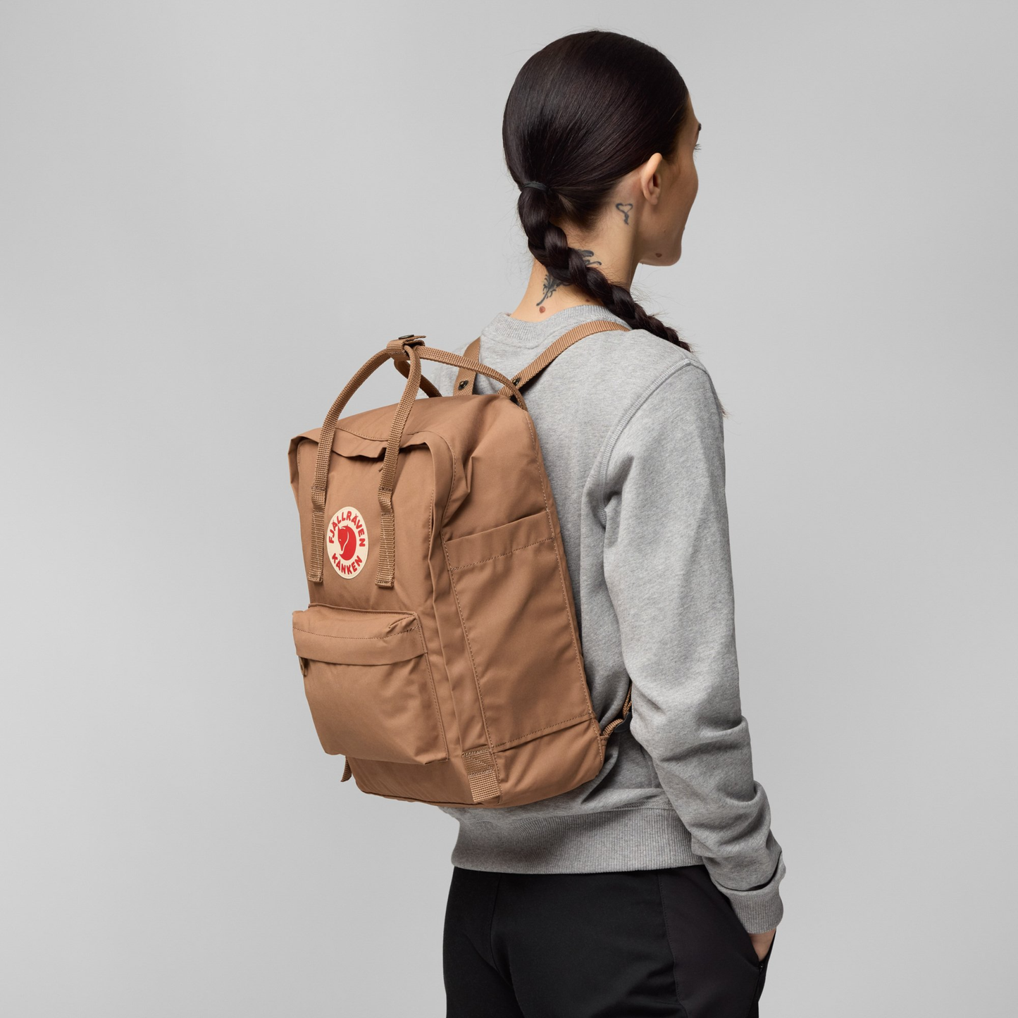 Side view of woman wearing The Fjallraven Kanken Backpack which is made from hard-wearing Vinylon F fabric and features a spacious main compartment with a full-length zip, removable PE foam seat cushion, two flat side pockets, a zipped front pocket, adjustable shoulder straps, and top handles. Durable and practical backpack for everyday use.