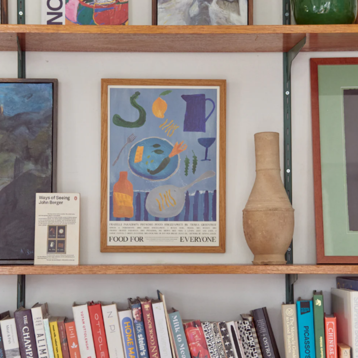 Shown on a book shelf Food for Everyone Fratelli Paradiso Pistachio pesto strozzapreti Poster in calming blue tones by Gemma Leslie, inspired by Trisha Greentree's Pistachio Pesto Strozzapreti recipe; printed on recycled coffee cup paper, unframed