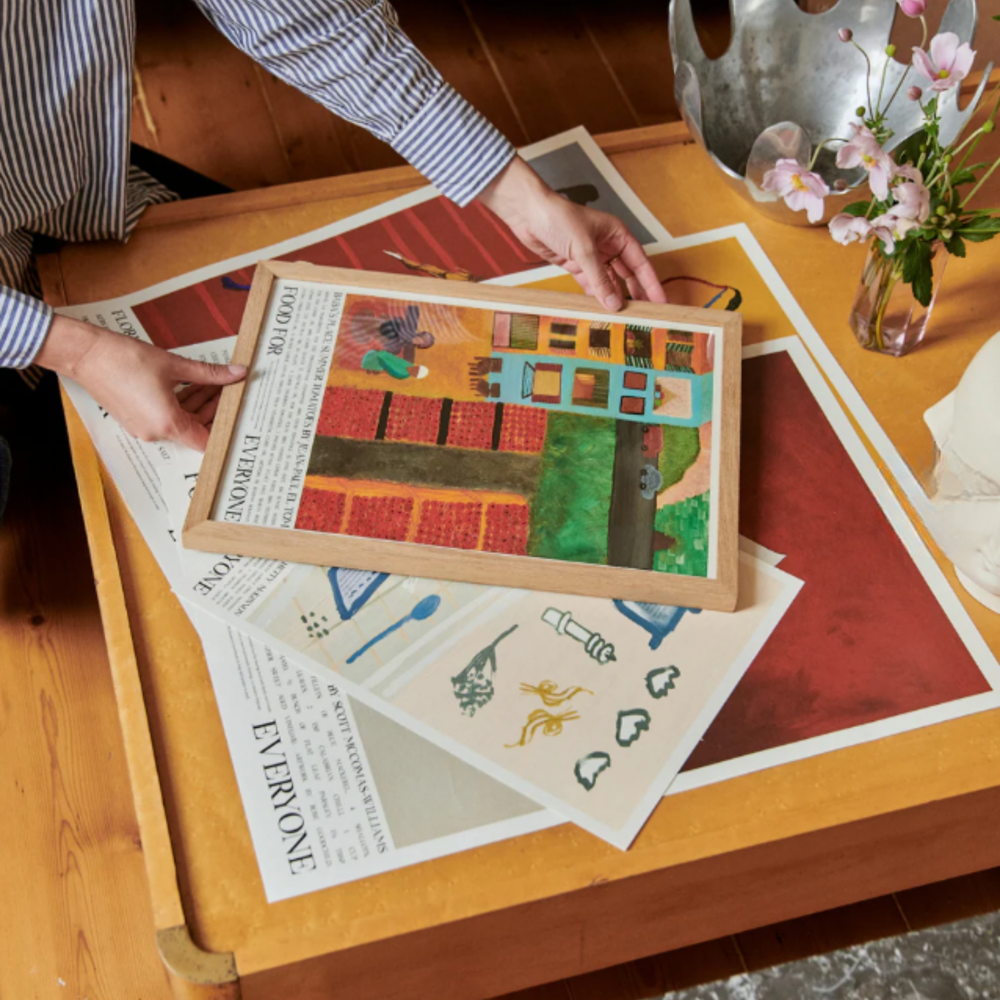 On a table being held the Food for Everyone Limited Edition Baba's Place Poster A2 printed on recycled coffee cup paper, featuring Rahma Soliman's warm summer illustration inspired by chef Jean-Paul El Tom's tomato recipe; unframed