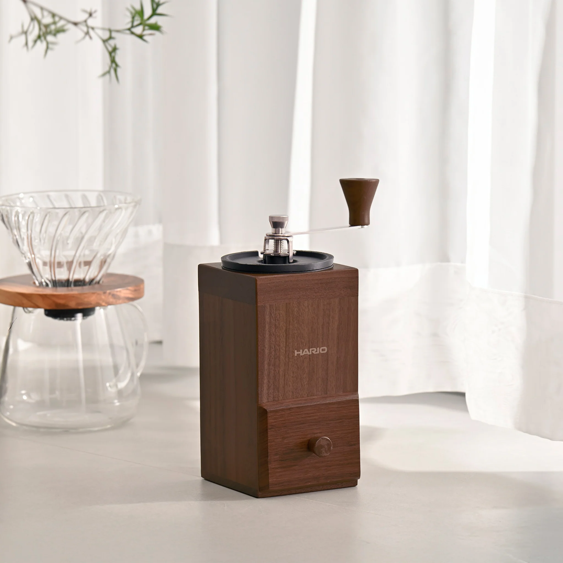 Seen on a table, the Hario Coffee Mill Cube 3-4 cups wooden coffee grinder with ceramic burr and stainless steel handle.