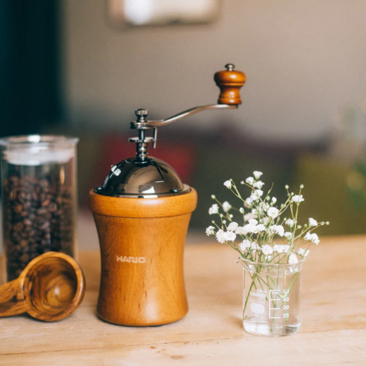 Table view with coffee and flowers of Hario Dome Coffee Mill, natural wood finish with rounded body and tapered center for easy grip, featuring a hand crank with wood handle and ceramic burr for consistent manual grinding. Compact and rustic design, ideal for 35g of coffee grounds.