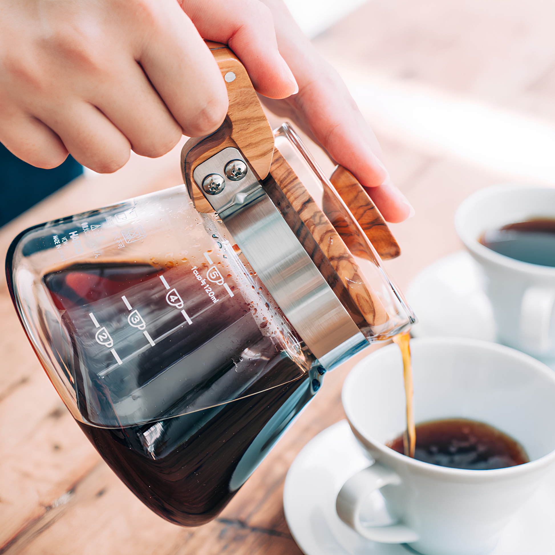 Pouring made coffee from the Hario Glass Coffee Server 600 ml Olive Wood which has pot from heatproof glass, lid from olive wood, handle, band and screw from stainless steel, with brass screw, featuring natural wood making each piece unique.