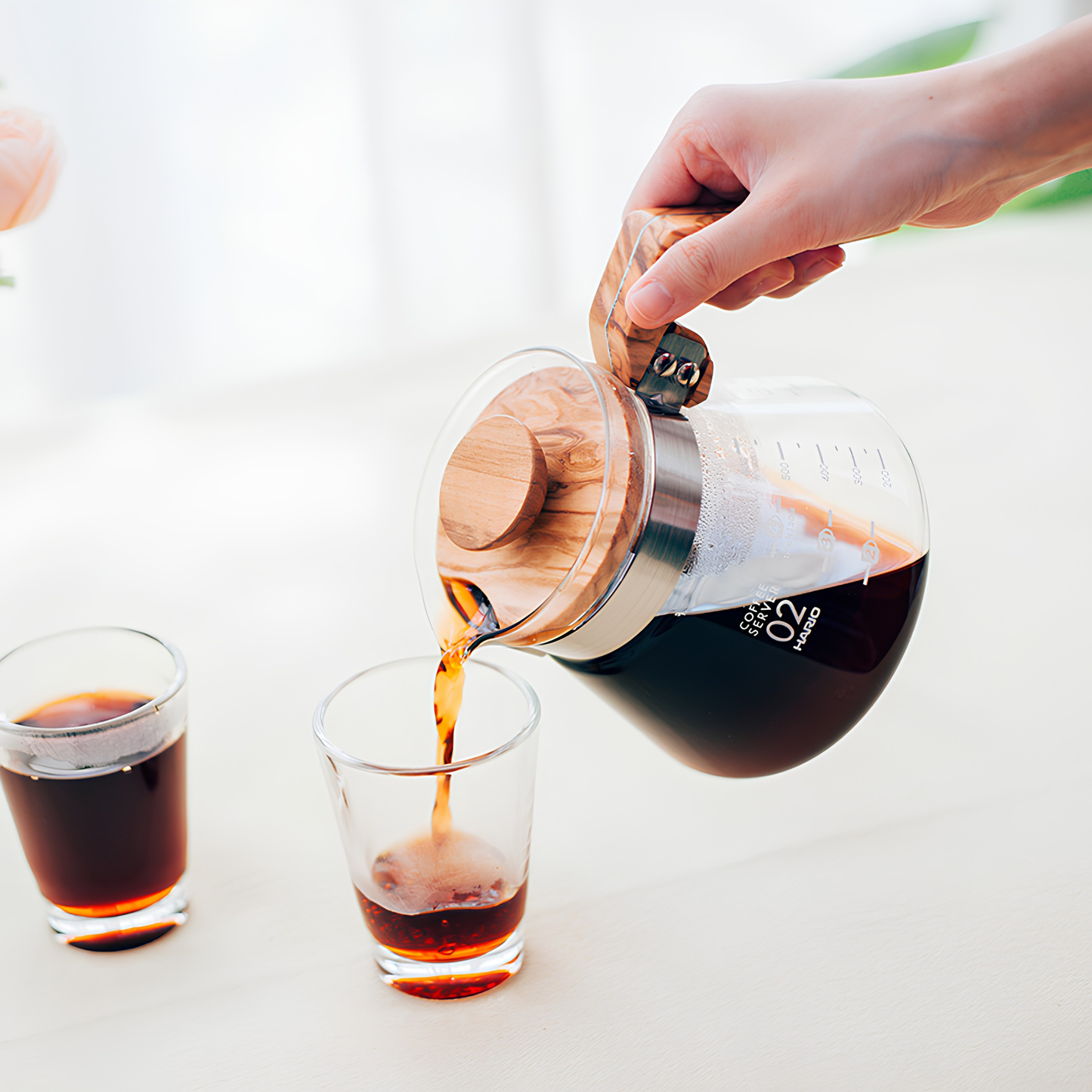 Another image of pouring coffee to the glass from the Hario Glass Coffee Server 600 ml Olive Wood which has pot from heatproof glass, lid from olive wood, handle, band and screw from stainless steel, with brass screw, featuring natural wood making each piece unique.