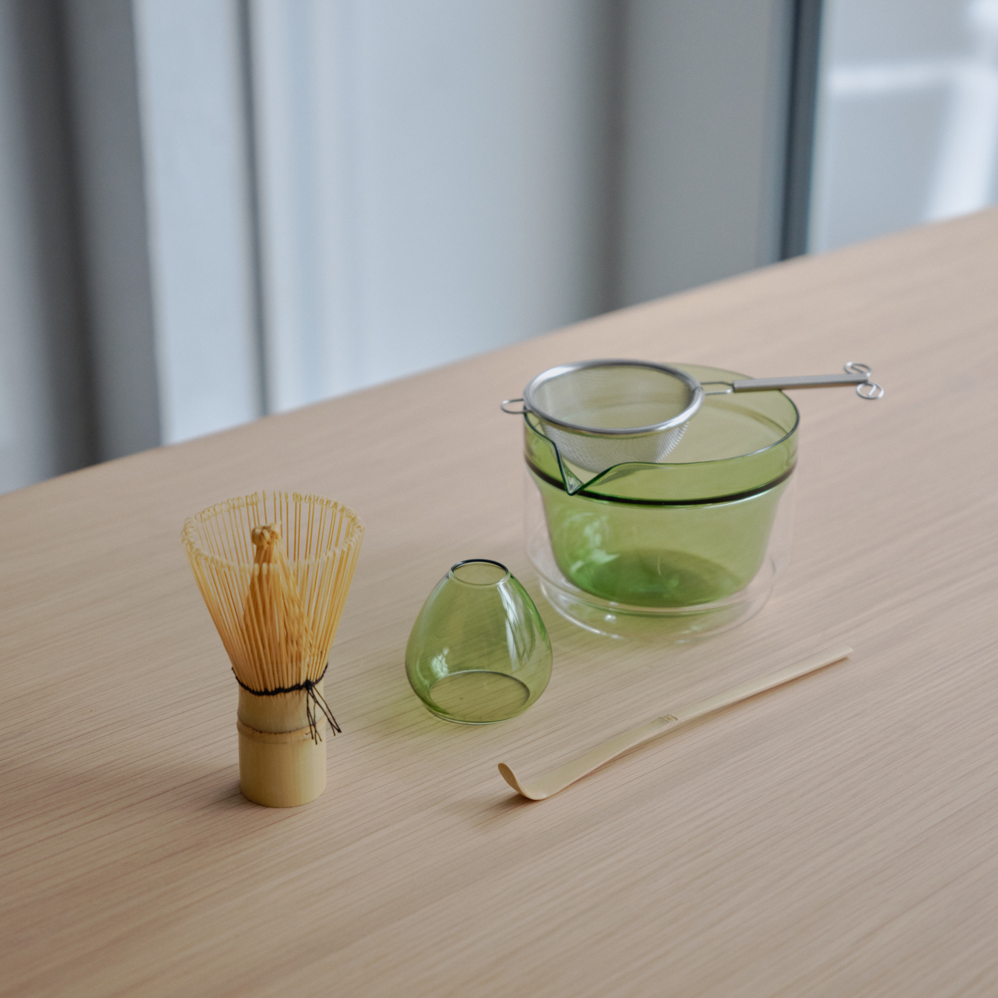 Seen on the table the items from the Hario Matcha Tea Set Green which includes a Double-walled Matcha Bowl, stand from heatproof glass, Chasen or the whisk, chashaku or measuring spoon from bamboo and tea sifter from stainless steel.