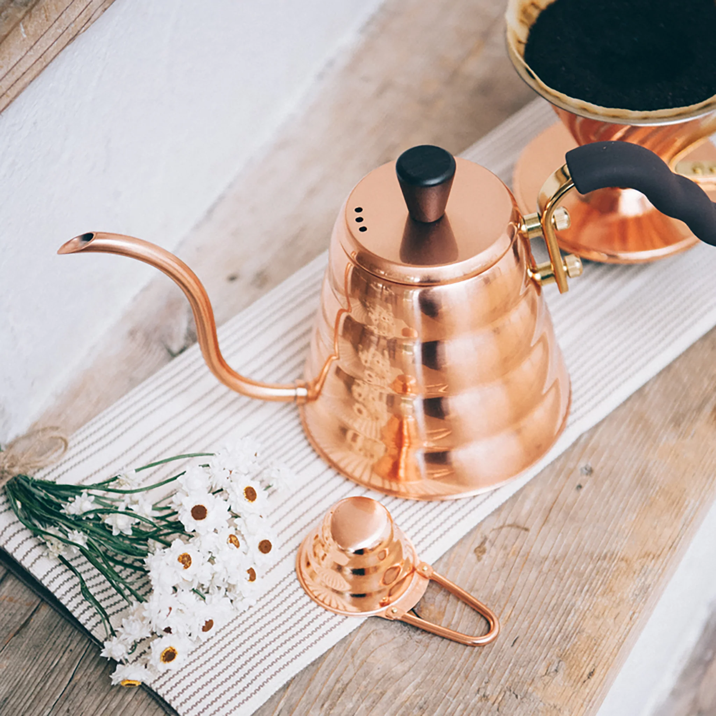 On a cloth on wooden surface, the Hario V60 Drip Kettle Buono 700ml Copper which includes Body of kettle and lid from copper, nickel and sealed with baked coating, lid knob from natural wood, handle from brass and handle cover from silicone rubber, luxurious of all kettles, iconic shape.
