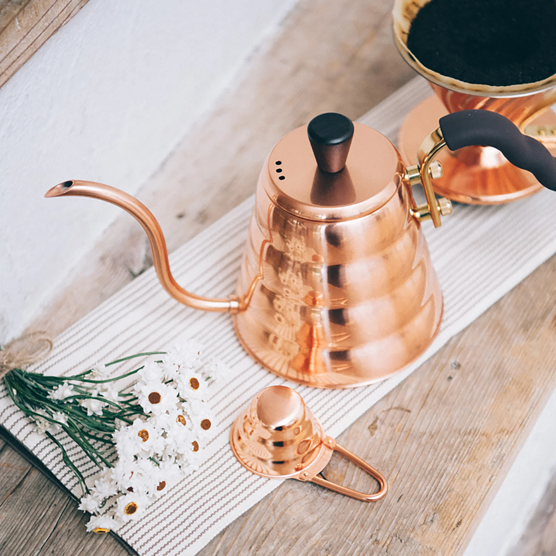 On a cloth on wooden surface, the Hario V60 Drip Kettle Buono 700ml Copper which includes Body of kettle and lid from copper, nickel and sealed with baked coating, lid knob from natural wood, handle from brass and handle cover from silicone rubber, luxurious of all kettles, iconic shape.