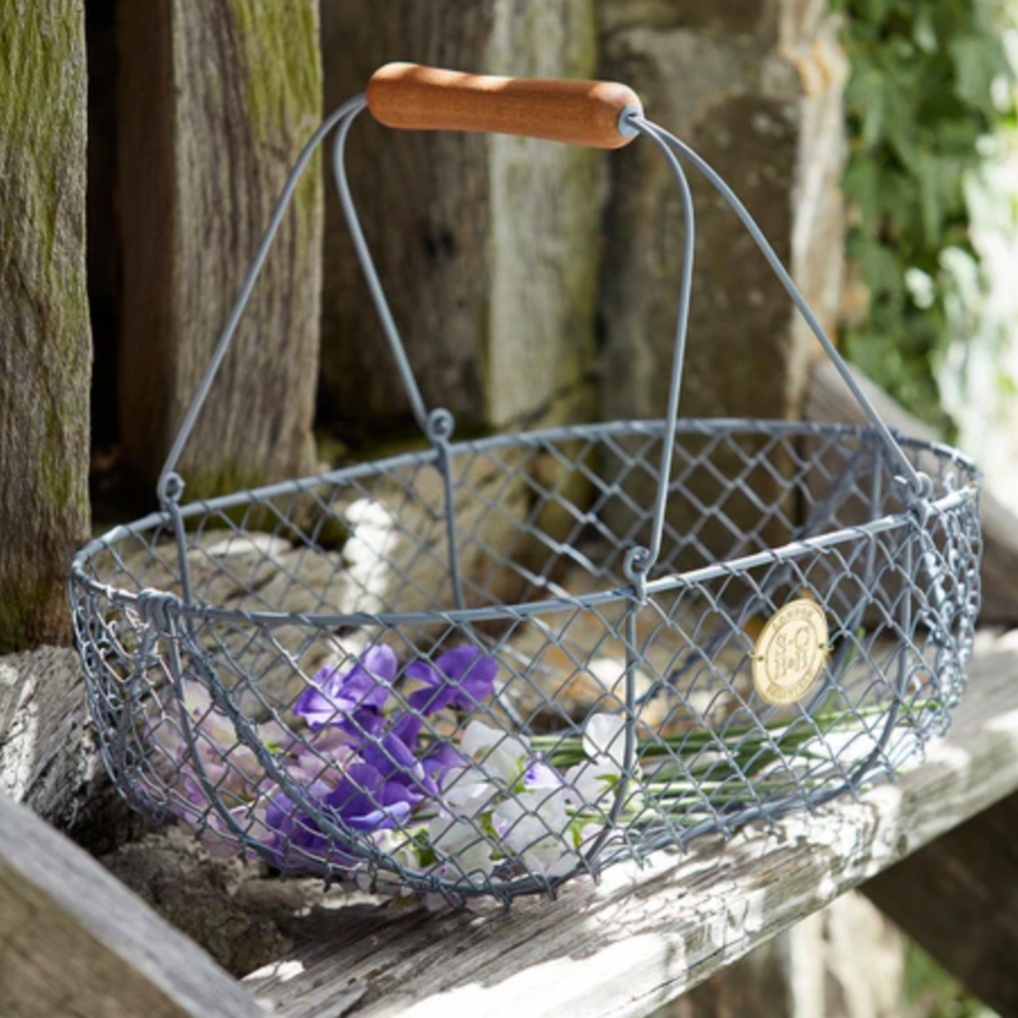 Burgon & Ball wire grey harvesting basket next to wooden fence with flowers in basket