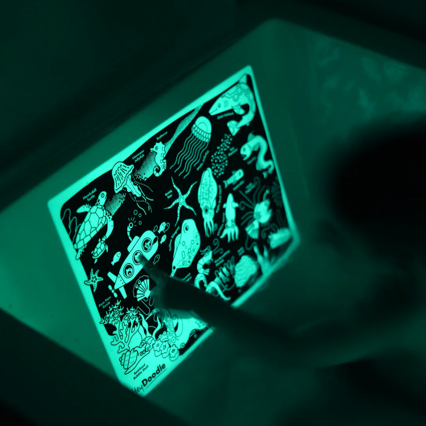 Image shown is glowing HeyDoodle Glow-in-the-Deep Mini Colouring & Doodle Mat - reusable silicone glow-in-the-dark travel mat with ocean creatures design, includes 3 non-toxic markers and pouch for mess-free creative fun on the go.