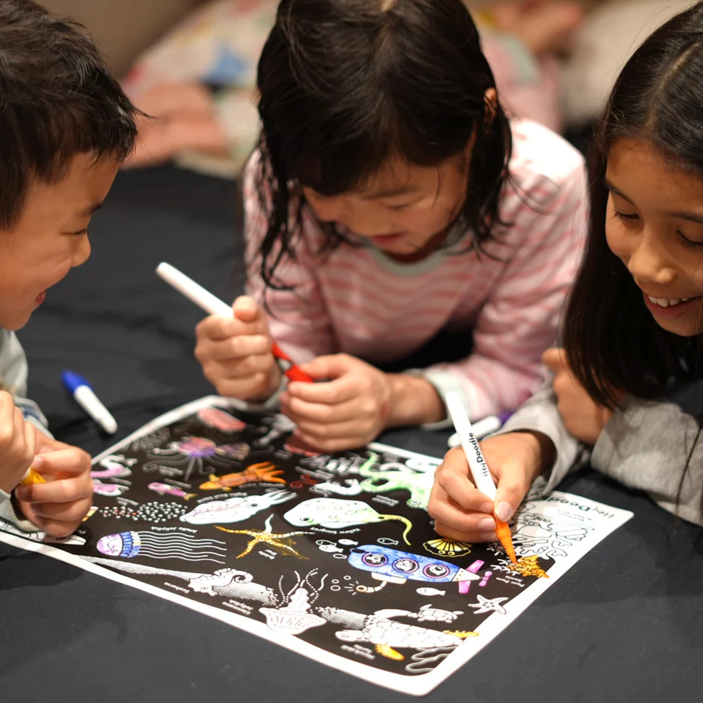 Kids coloring the HeyDoodle Glow-in-the-Deep Mini Colouring & Doodle Mat - reusable silicone glow-in-the-dark travel mat with ocean creatures design, includes 3 non-toxic markers and pouch for mess-free creative fun on the go.