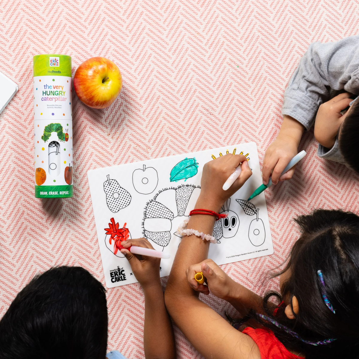 Three children color a The Very Hungry Caterpillar sheet with markers beside the Hey Doodle Mini Colouring & Doodle Mat.