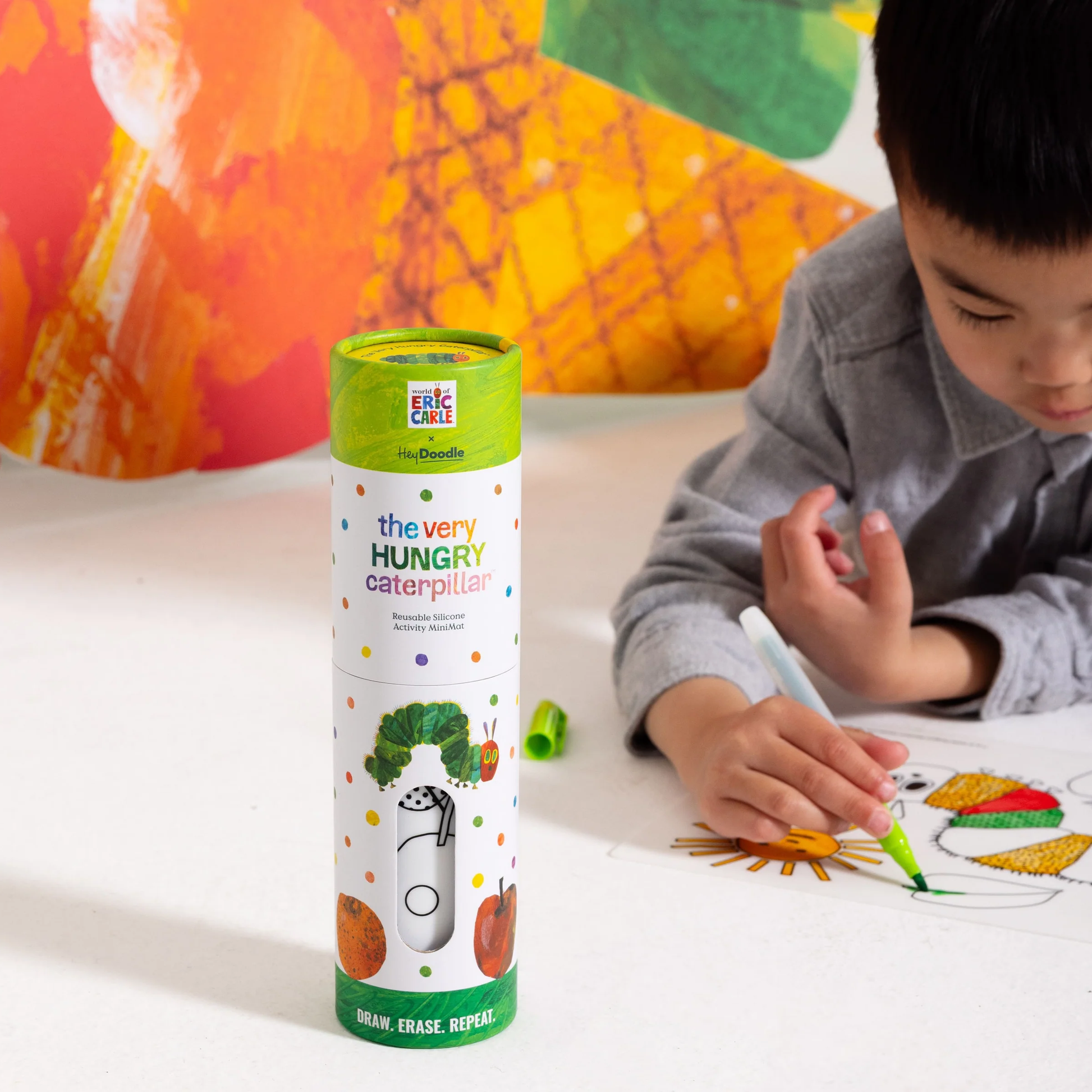 A child colors a drawing with markers beside a Hey Doodle Mini Colouring & Doodle Mat The Very Hungry Caterpillar tube.