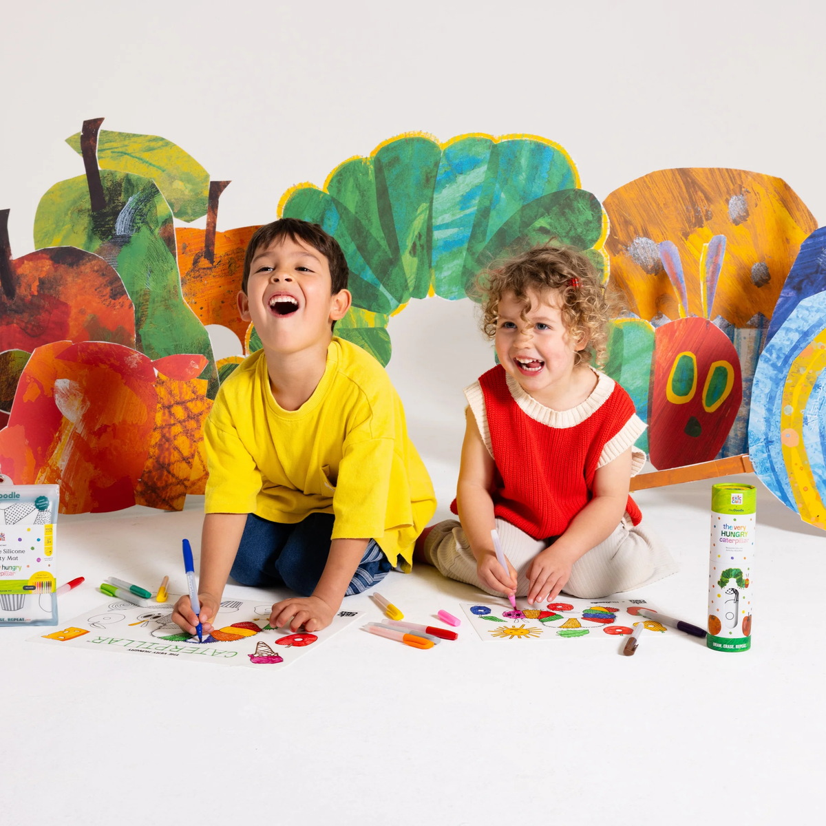 Two children smile and color on a Hey Doodle Mini Colouring & Doodle Mat inspired by The Very Hungry Caterpillar.