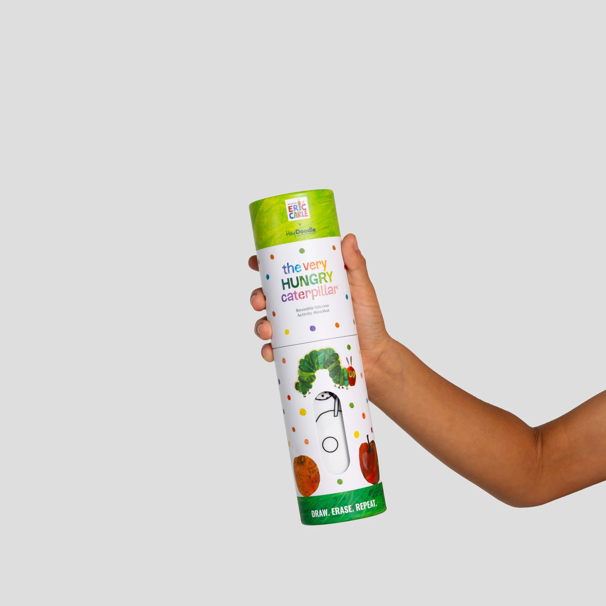 A child's hand holds the Hey Doodle Mini Colouring & Doodle Mat The Very Hungry Caterpillar tube with dots and fruit designs.