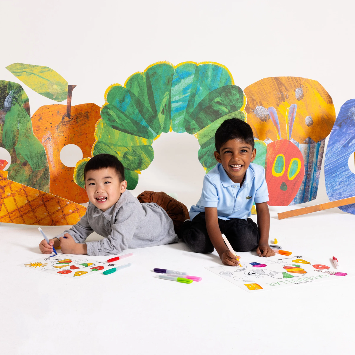 Two young boys smile as they color on paper, with the Hey Doodle Mini Colouring & Doodle Mat with the life size images behind them.