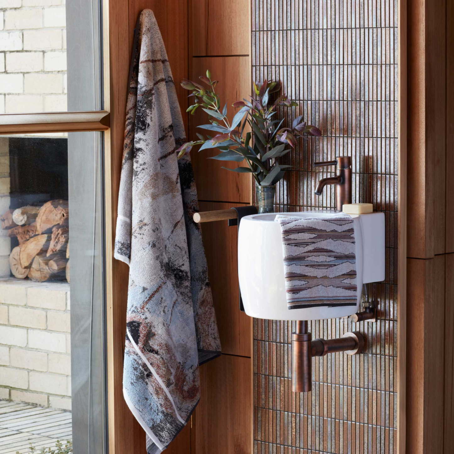 Shown being hung on the wall beside the window, Innate NT Morning Bath Sheet 100cm x 150cm bath sheet in warm earthy tones inspired by Australia's Top End landscape, made from 100% OEKO-TEX® certified cotton, featuring an absorbent six-colour jacquard weave with rich texture and weight, responsibly woven in Portugal for everyday premium use.