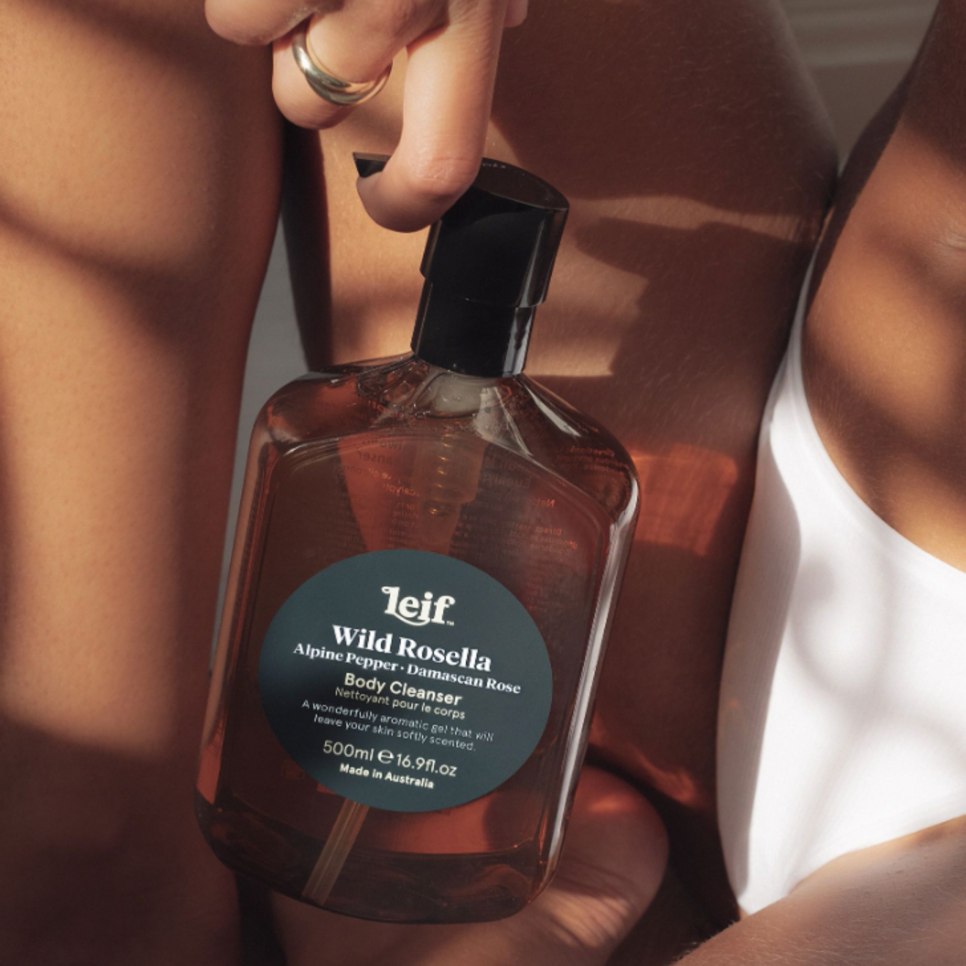 Woman posing in bathroom with Leif body cleanser Wild Rosella 500ml between her legs and arms. 