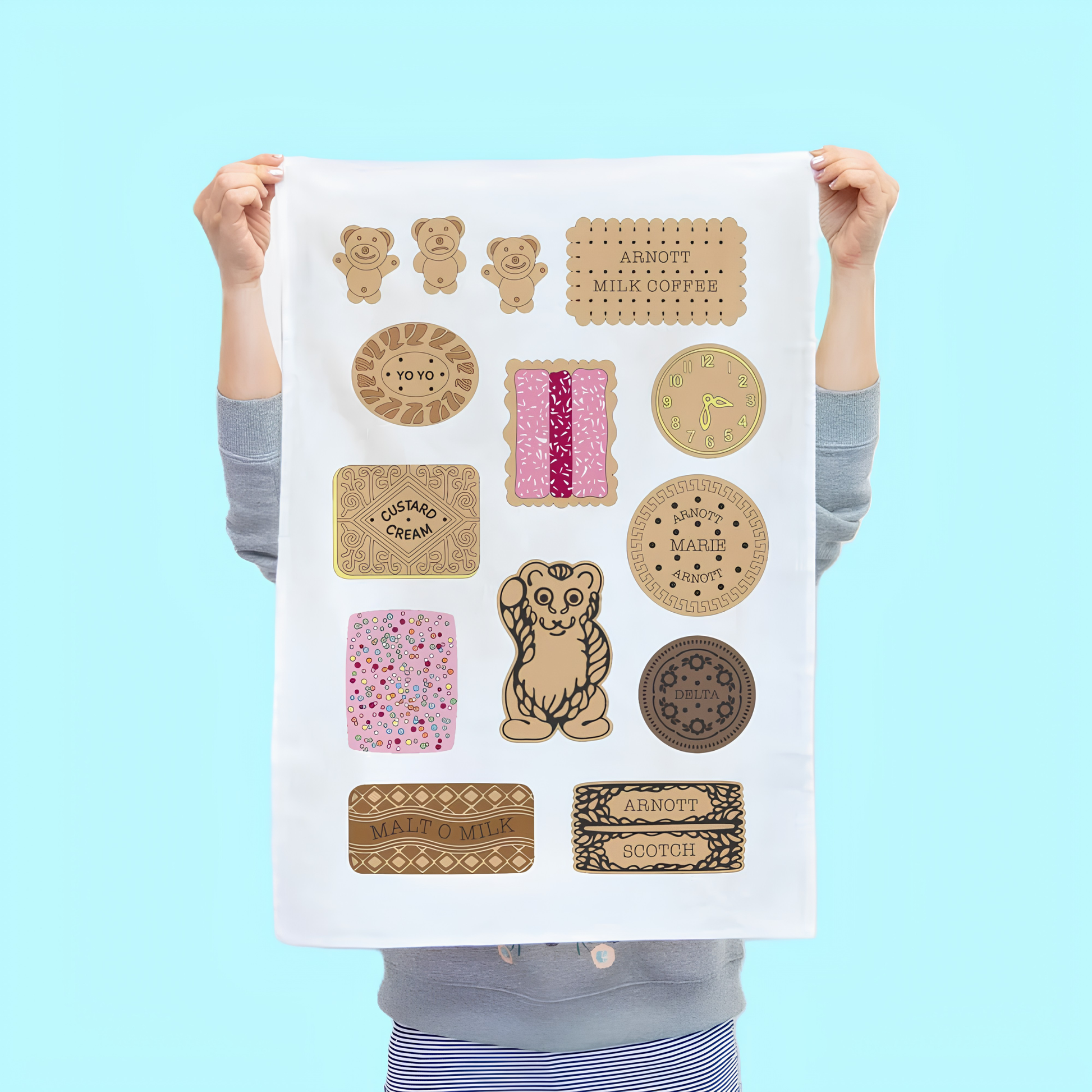 Held and raised by a girl the front of the Make Me Iconic Tea Towel Bickies, Australia's most iconic biscuits, Delta Creams, Malt-O-Milk, Teddy Bear and more put on a tea towel. Made from high quality, absorbent fabric.