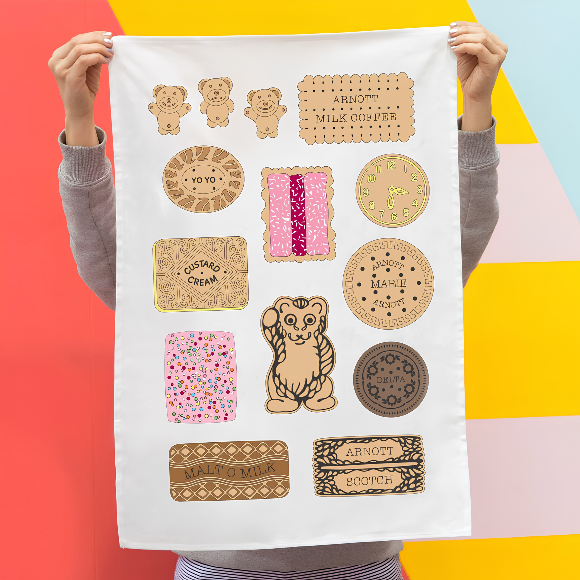 Front view of the Make Me Iconic Bickies Tea Towel held up by a girl against a colorful background, featuring Australia's iconic biscuits like Delta Creams, Malt-O-Milk, and Teddy Bear. Made from high-quality, absorbent fabric.
