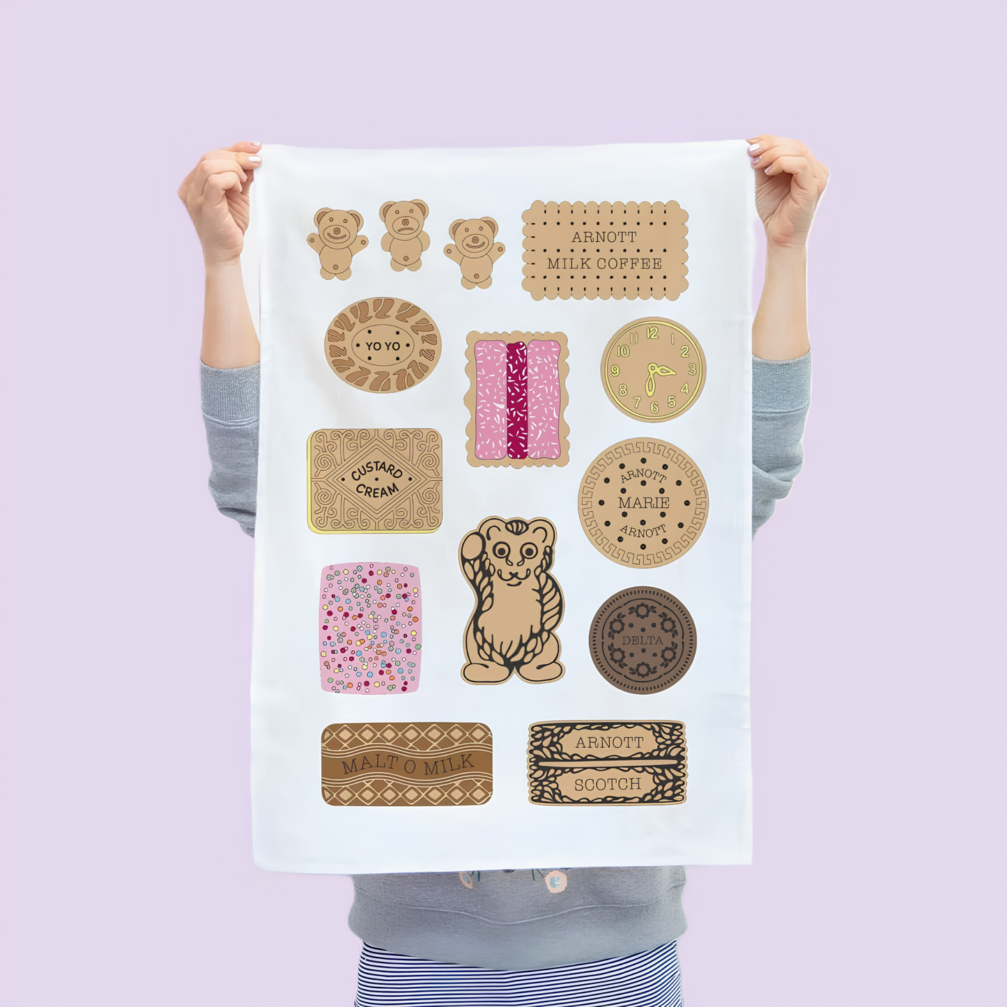 Another front view of the Make Me Iconic Bickies Tea Towel held up by a girl against a light purple background, featuring Australia's iconic biscuits like Delta Creams, Malt-O-Milk, and Teddy Bear. Made from high-quality, absorbent fabric.