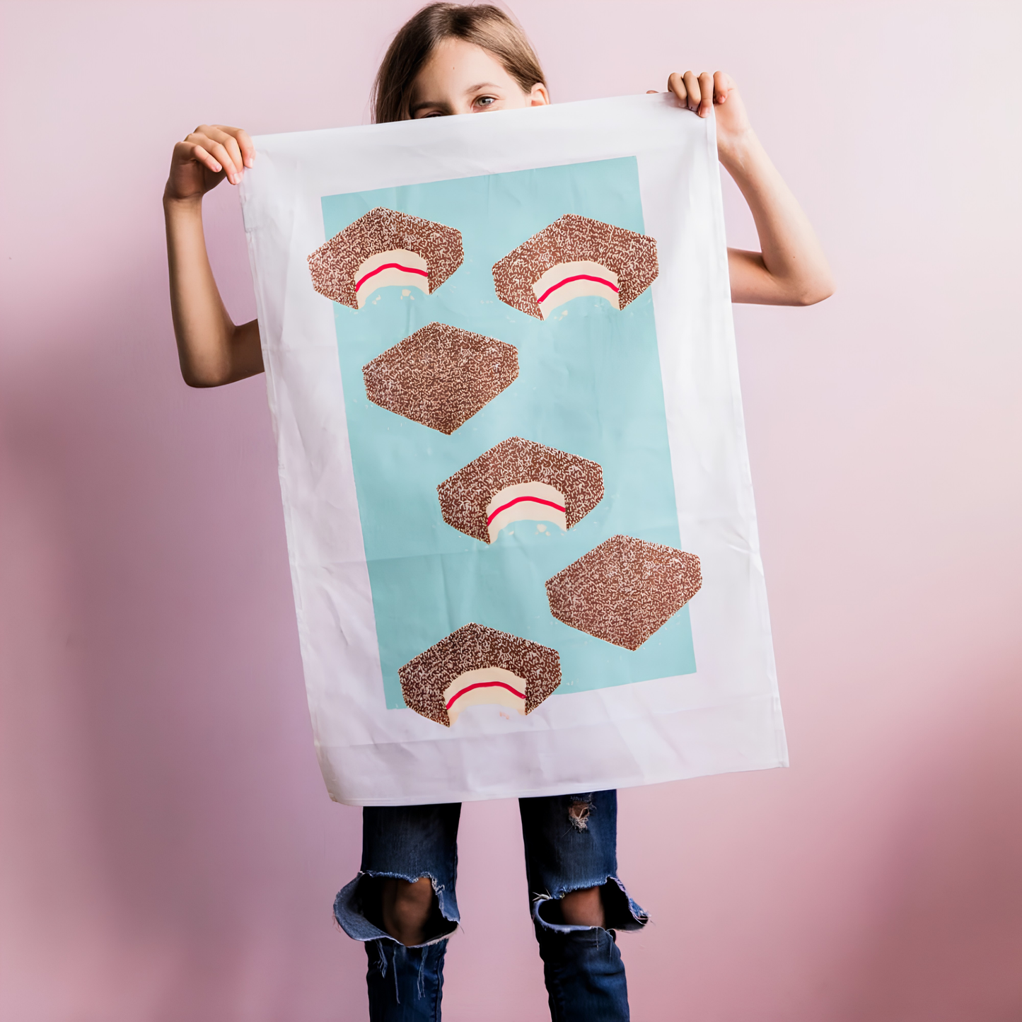 A girl holding the front of the Make Me Iconic Lamington Tea Towel featuring Australia's classic chocolate-coated, coconut-covered treat. Made from high-quality, absorbent fabric.
