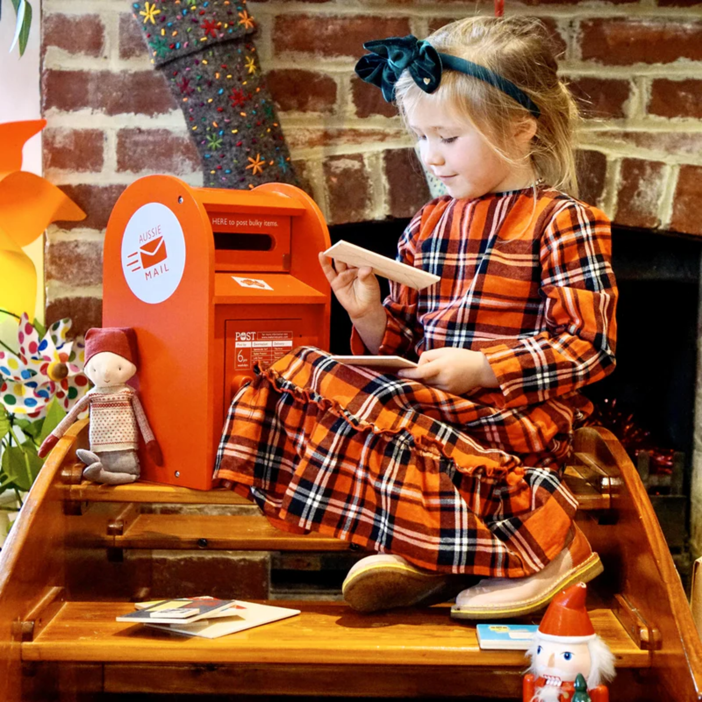 Showing a little girl reading a wooden letter from the Make Me Iconic wooden red post box toy set featuring a working pull-down slot, six mix-and-match Velcro stamps, and six wooden letters and postcards - a nostalgic, eco-friendly playset designed in Australia to inspire creativity and role play for children aged 3 and up.
