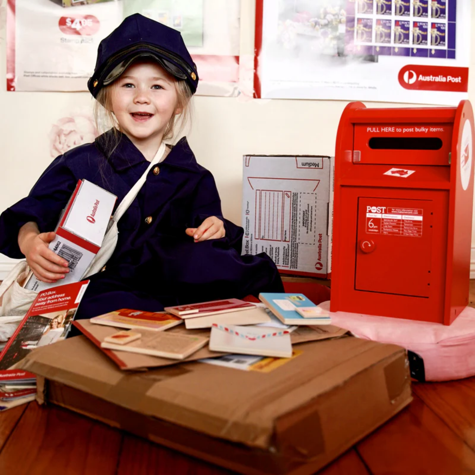 Showing a mail little girl with the Make Me Iconic wooden red post box toy set featuring a working pull-down slot, six mix-and-match Velcro stamps, and six wooden letters and postcards - a nostalgic, eco-friendly playset designed in Australia to inspire creativity and role play for children aged 3 and up.