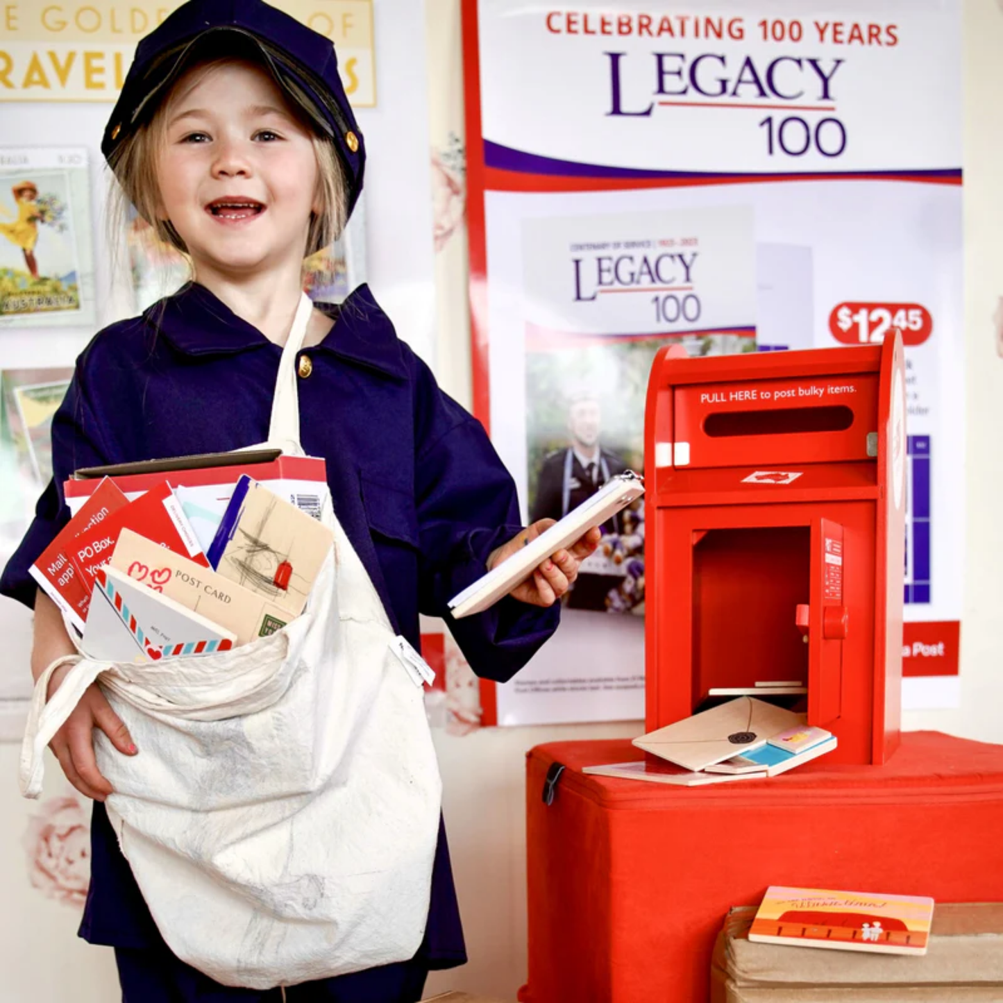 Showing a mail little girl carrying wooden cards with the Make Me Iconic wooden red post box toy set featuring a working pull-down slot, six mix-and-match Velcro stamps, and six wooden letters and postcards - a nostalgic, eco-friendly playset designed in Australia to inspire creativity and role play for children aged 3 and up.
