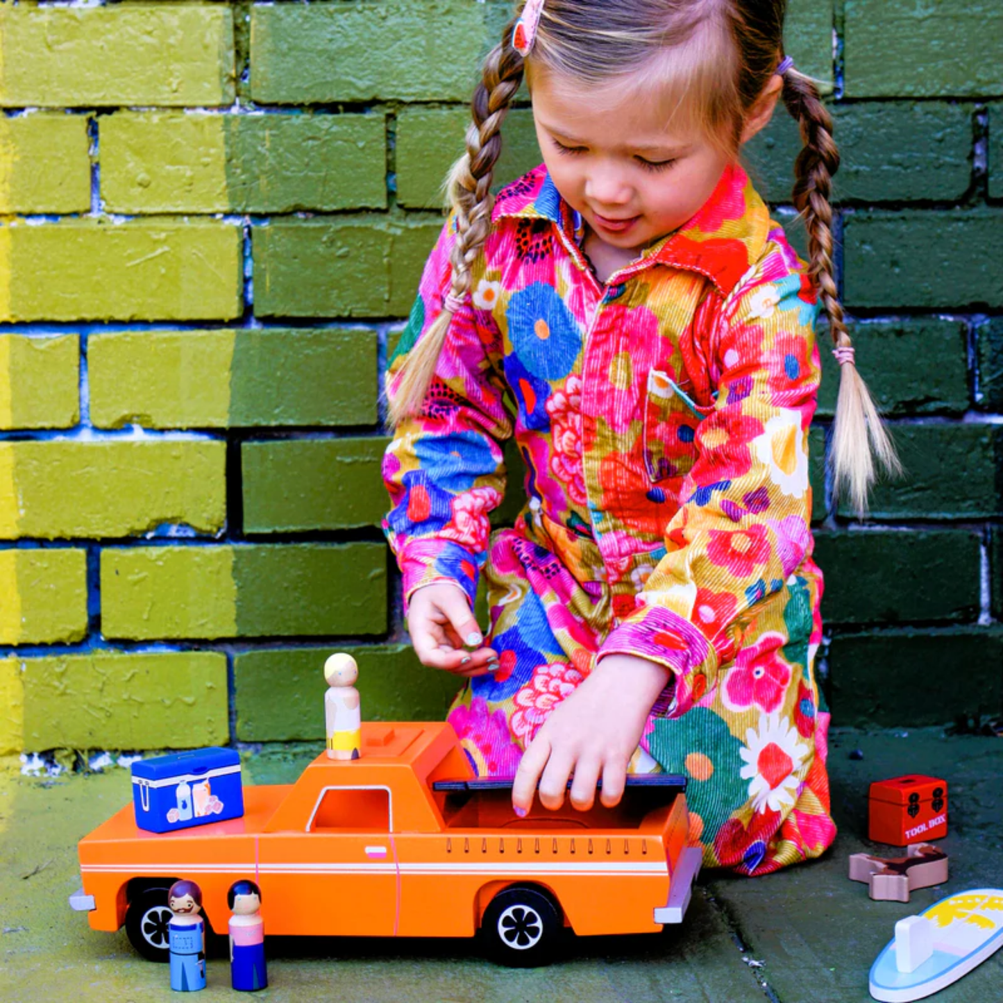 Showing a girl playing the Make Me Iconic wooden blue Ute toy set with rolling wheels, lift-off roof, three wooden passengers, a Kelpie dog, surfboard, esky, and toolbox - a nostalgic Aussie-inspired playset designed in Australia to spark creativity and hands-on imaginative fun for children aged 3 and up.