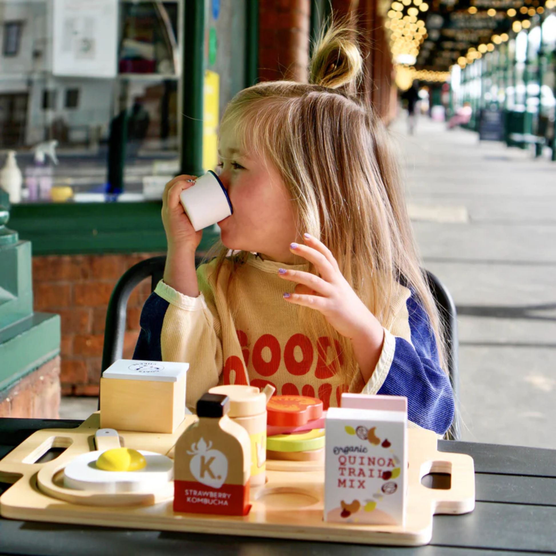 Little girl playing the Make Me Iconic wooden toy breakfast set - multicolor wood play set featuring 21 café-style pieces including avocado toast, kombucha, and sliceable goat's cheese for creative pretend play.