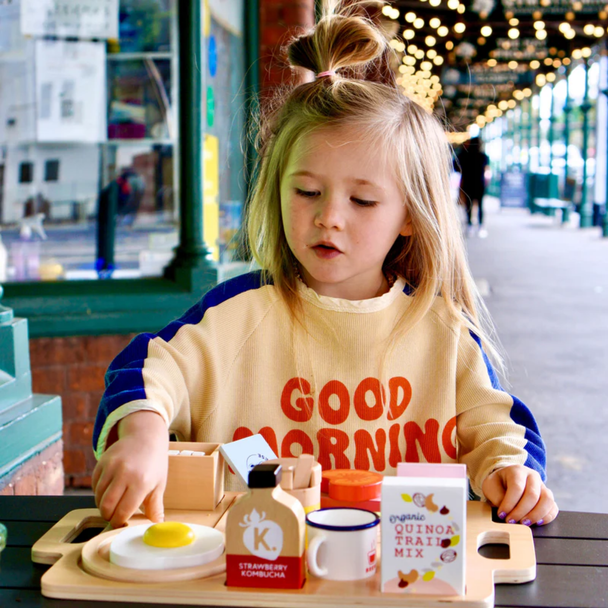 Little girl playing the Make Me Iconic wooden toy breakfast set - multicolor wood play set featuring 21 café-style pieces including avocado toast, kombucha, and sliceable goat's cheese for creative pretend play.