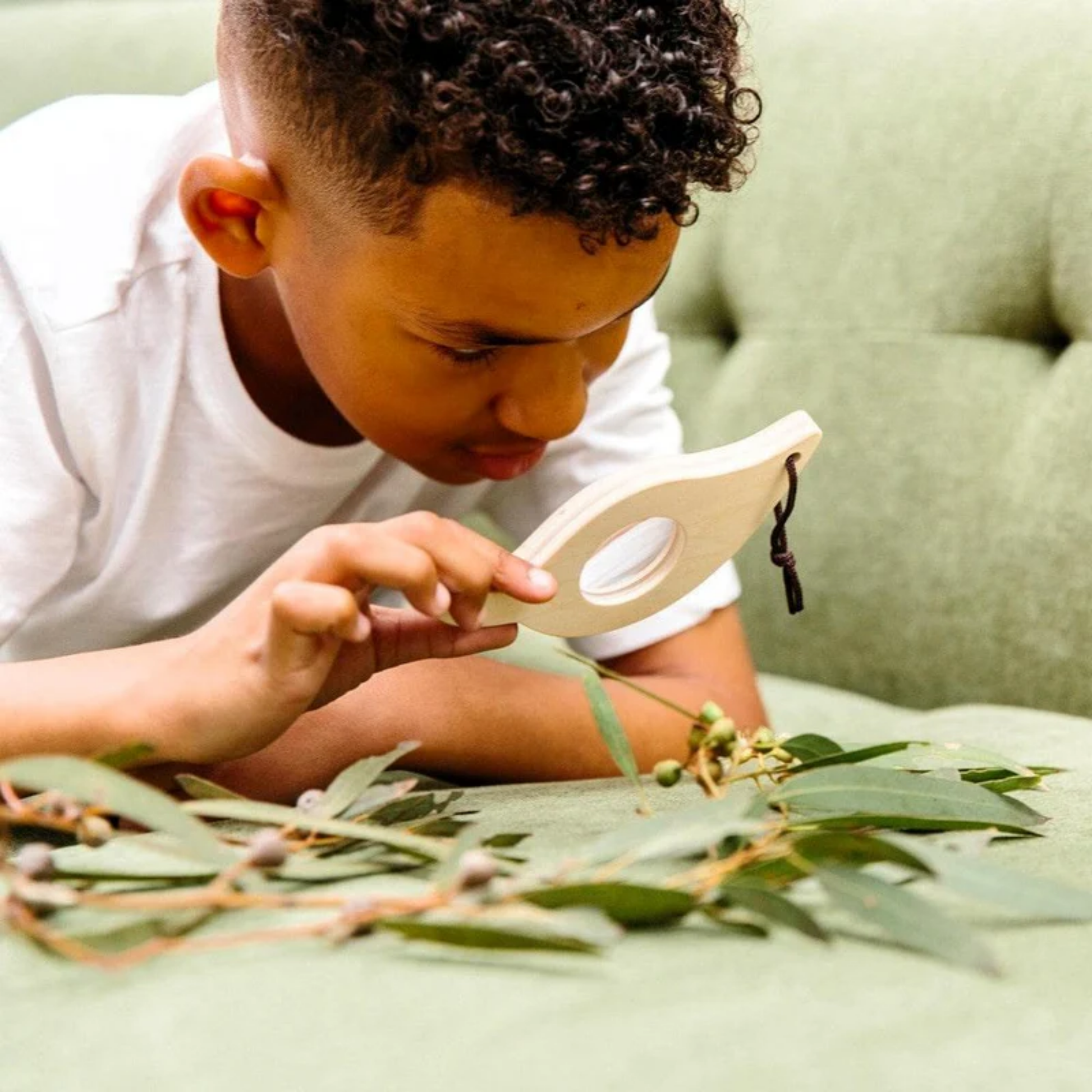 A boy magnifying a plant using Make Me Iconic Leaf Magnify Glass - Green wooden magnifying toy for kids, made from sturdy wood with a safe plastic lens and carry loop for outdoor exploration.