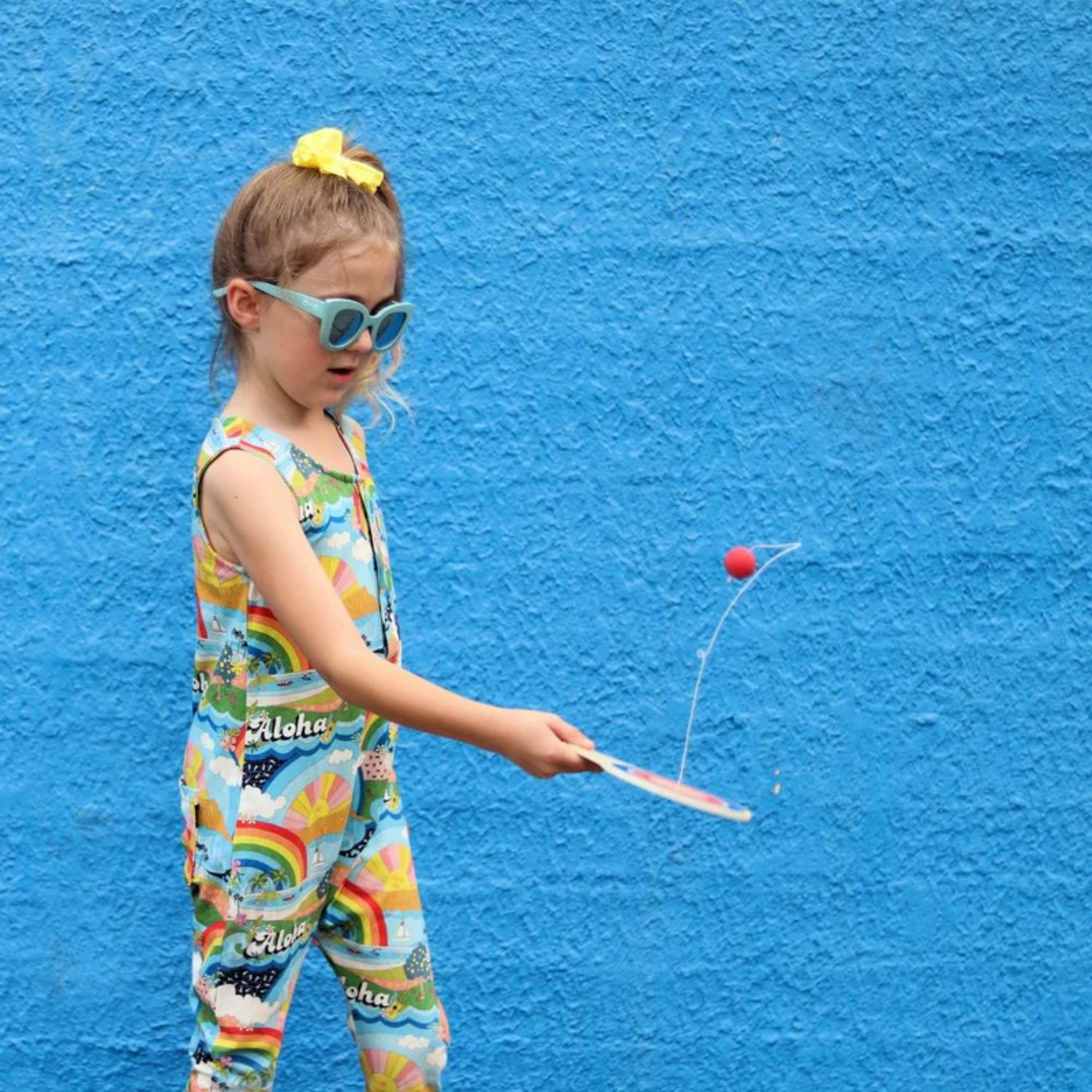 Little girl playing Make Me Iconic wooden paddle ball toy - natural plywood paddle with attached red rubber ball on elastic cord, a retro skill game that builds focus and coordination for kids and adults alike.