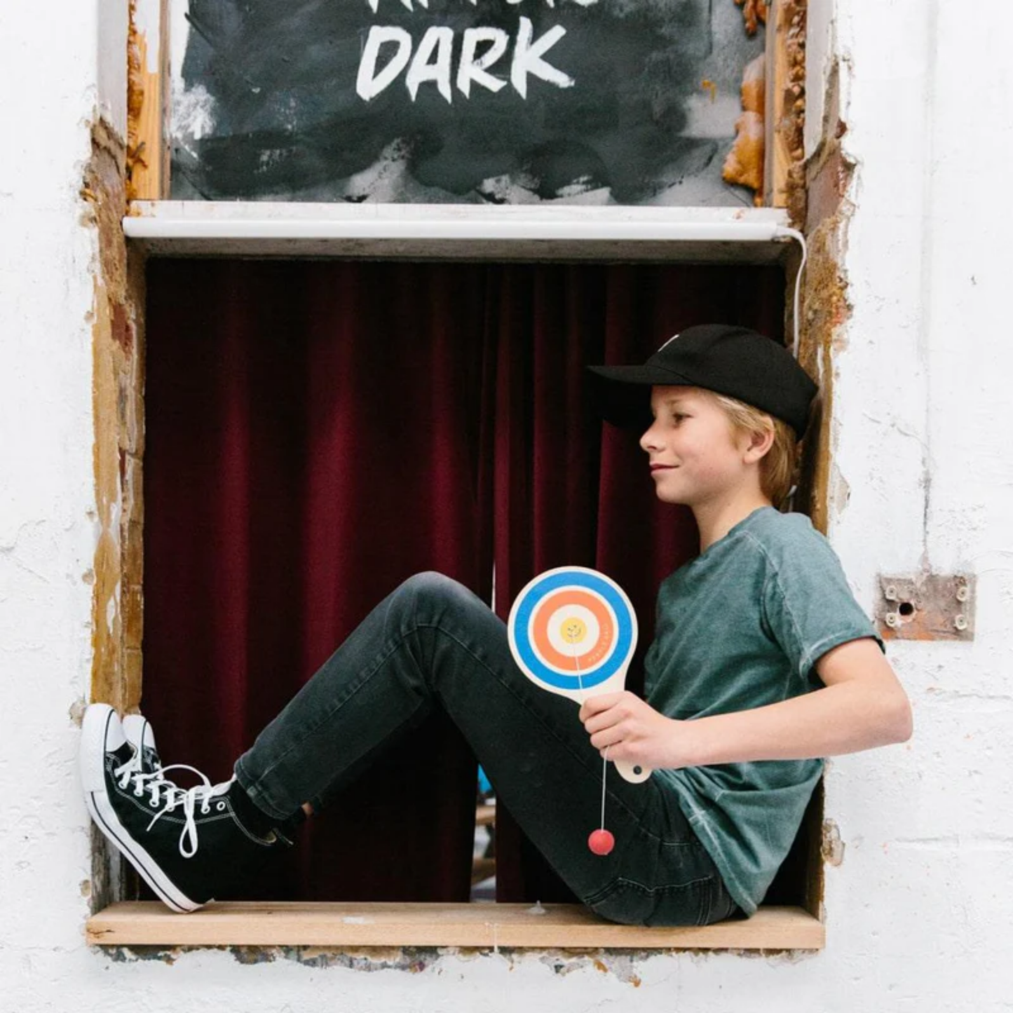 Shown being held by a boy Make Me Iconic wooden paddle ball toy - natural plywood paddle with attached red rubber ball on elastic cord, a retro skill game that builds focus and coordination for kids and adults alike.