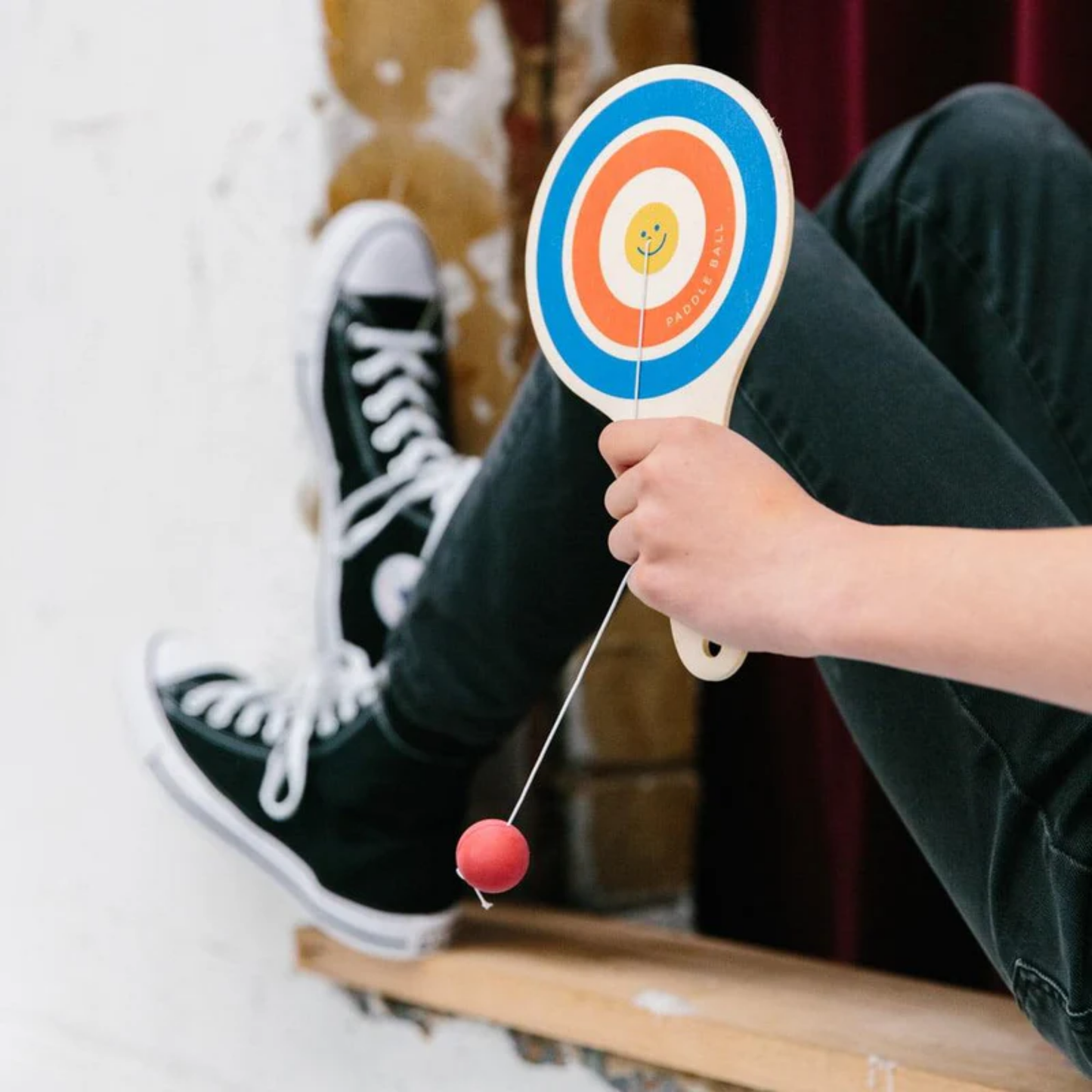 Upclose view being held by a boy Make Me Iconic wooden paddle ball toy - natural plywood paddle with attached red rubber ball on elastic cord, a retro skill game that builds focus and coordination for kids and adults alike.