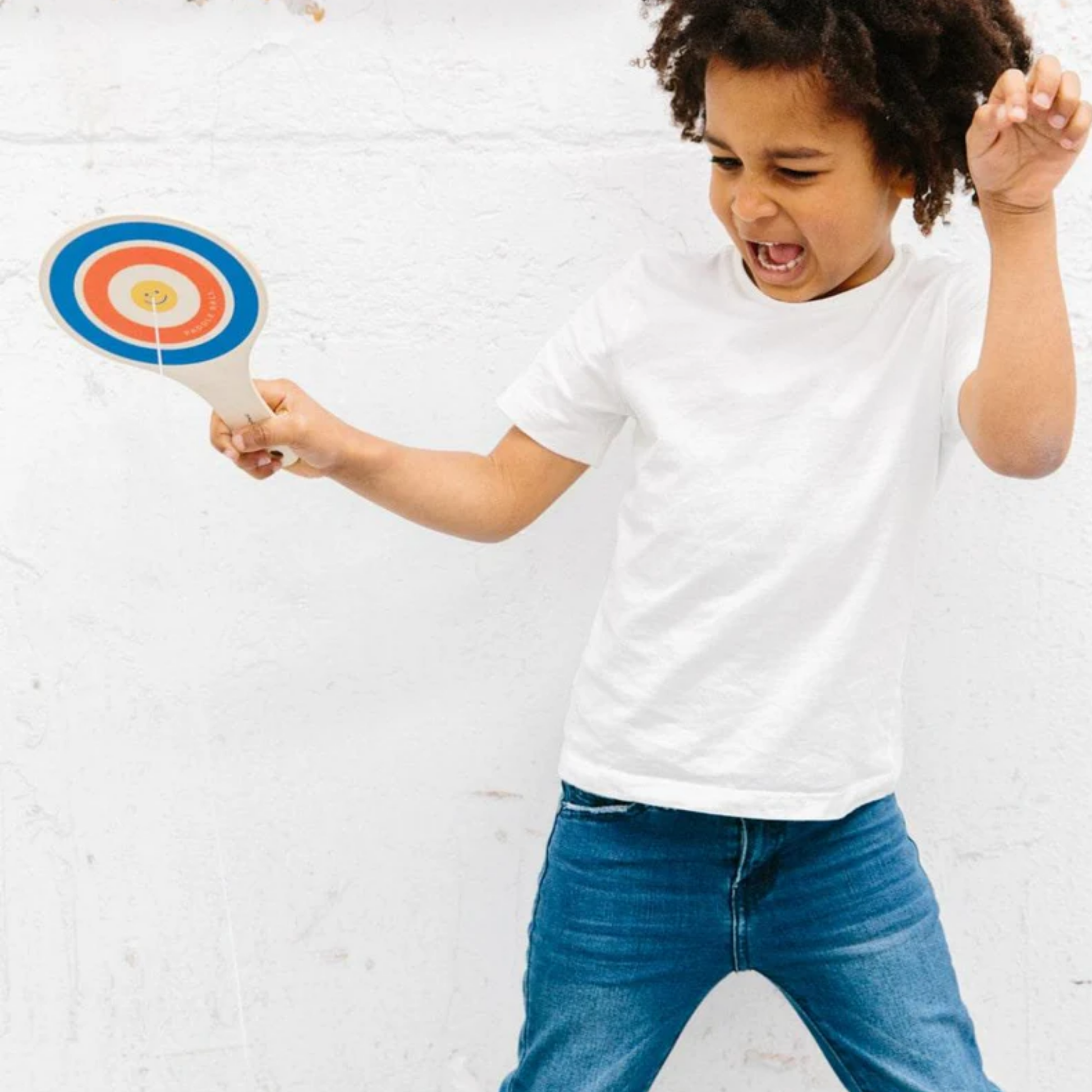 Little boy playing Make Me Iconic wooden paddle ball toy - natural plywood paddle with attached red rubber ball on elastic cord, a retro skill game that builds focus and coordination for kids and adults alike.