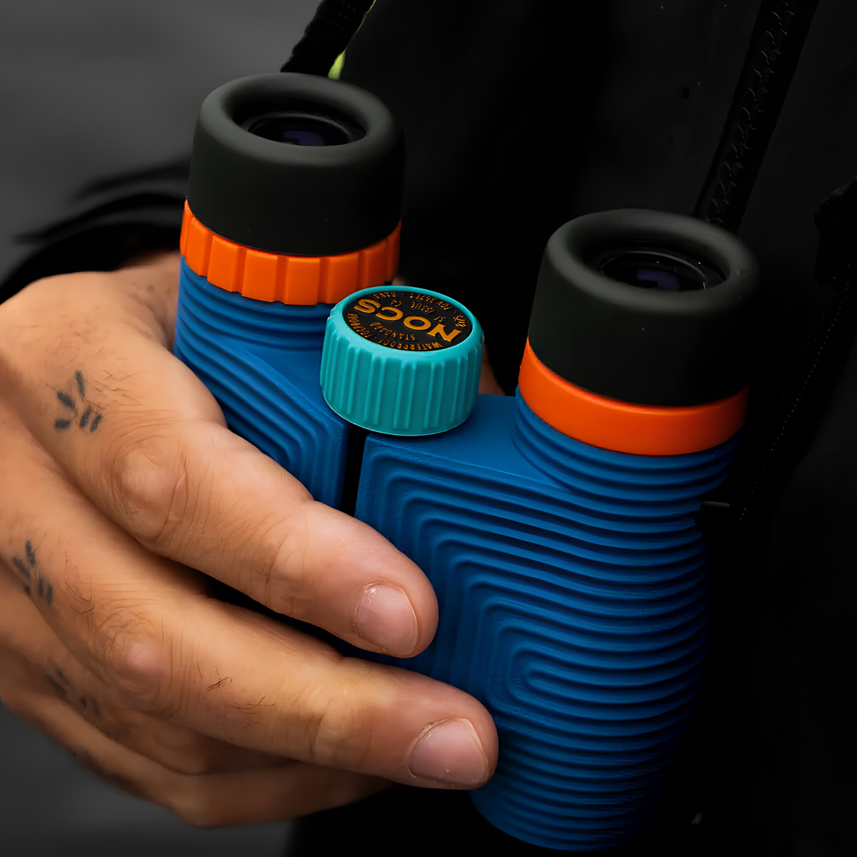 A hand holding the Nocs Provisions Binocular Standard Issue 8X25 Cobalt Blue that has multi-coated lenses and premium BaK4 prisms, waterproof, fogproof and compact.