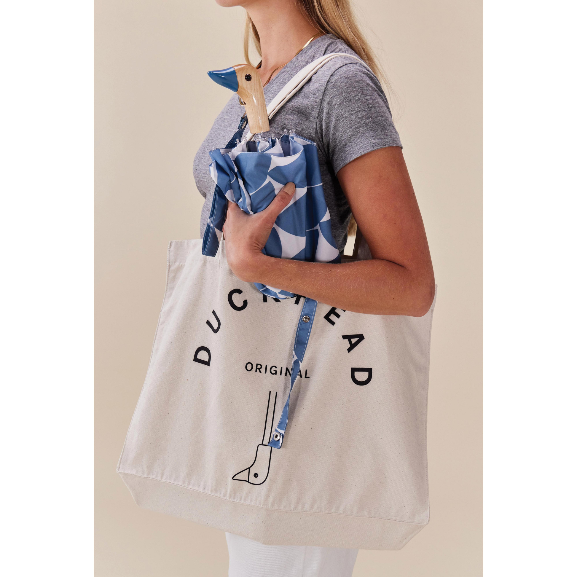 Side view of a woman holding the Folded inside its pouch, the Original Duckhead Duck Umbrella Compact Denim Moon from 100% recycled plastic bottles and robust to keep you dry, it fits into any backpack or tote bag, it is wind resistant, has automatic open button and high-strength steel frame. For everyday use no matter the weather.