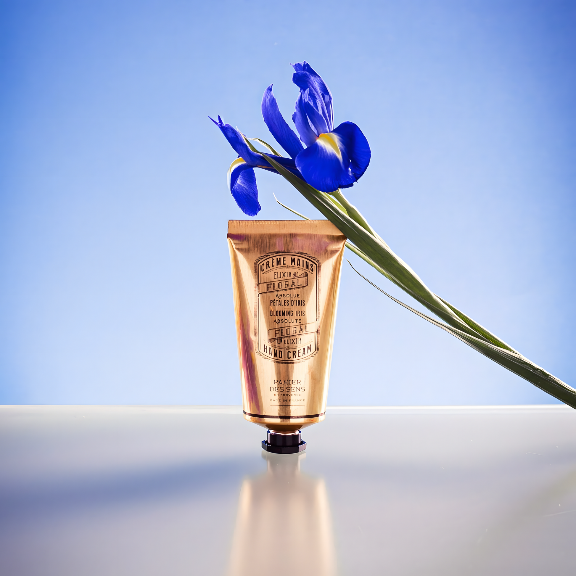 Fathom Blooming Iris hand cream 75ml standing shown with a flower leaning to it, on a surface with a blue background, it's a natural moisturizing hand lotion made with olive oil and iris, made in France.