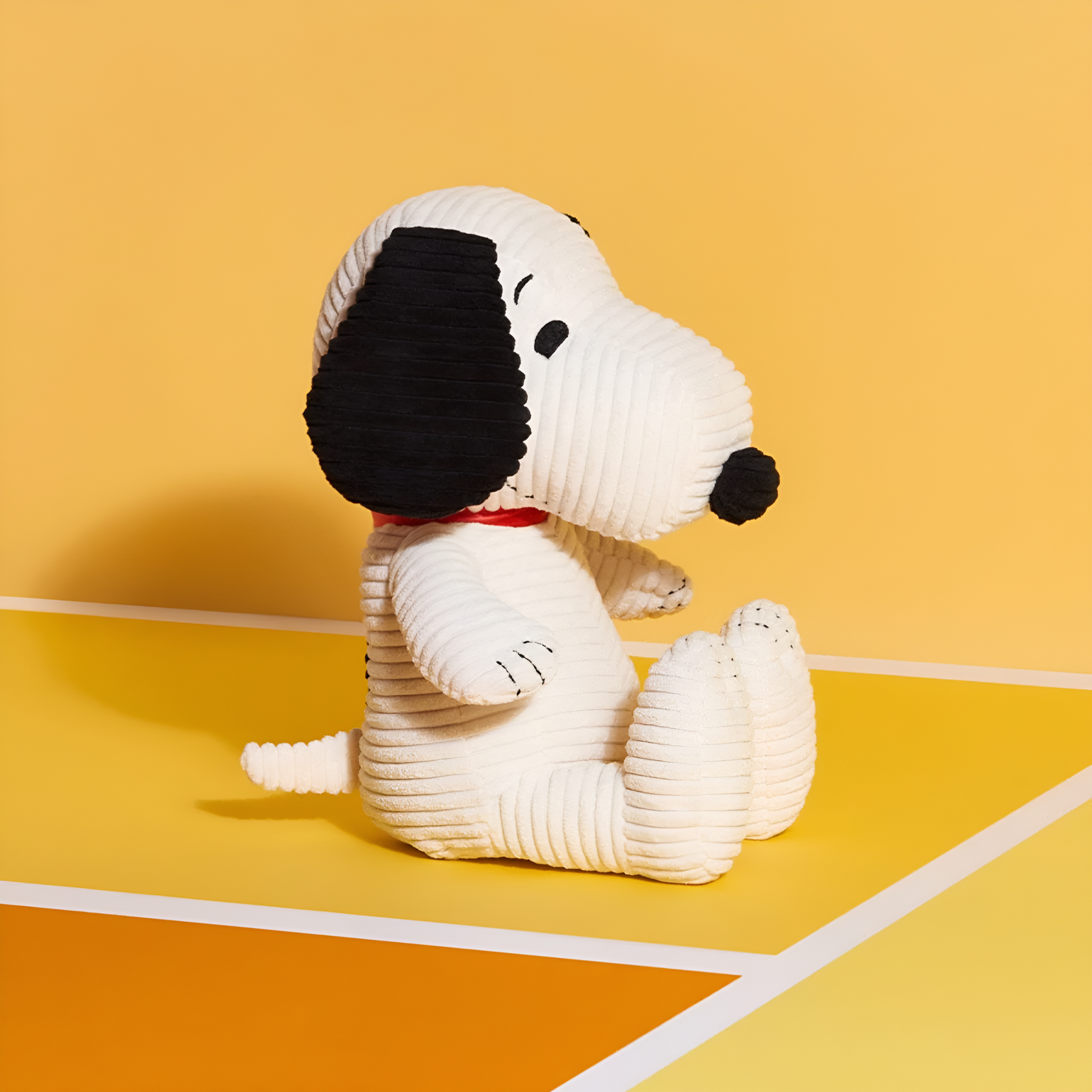 On a yellow surface the side angle of the Peanuts Snoopy Corduroy 27cm Cream, made from best corduroy, hand made and contains 100% recycled polyester, the world famous dog Snoopy can be paired with his little yellow friend Woodstuck.