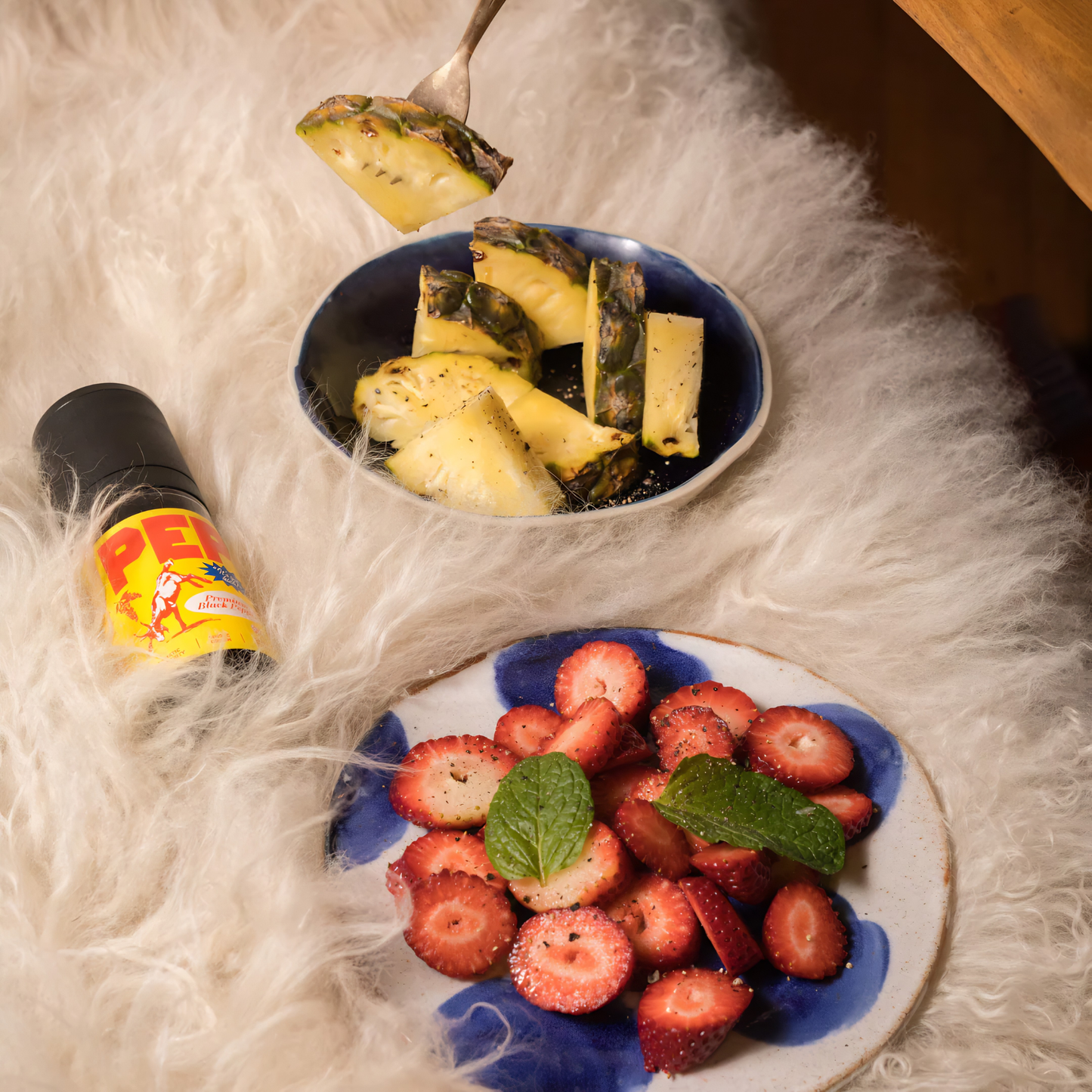 Laid on a fur like surface with plates of strawberries and pineapples, the PEP Pepper Grinder 50g, for fresh black peppercorns in a reusable smoky glass with yellow cover, with aromatic complexity grown in Cambodia.