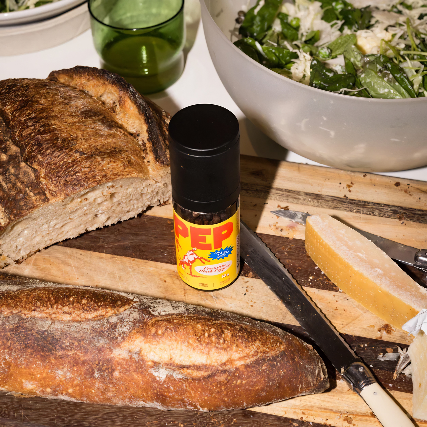 On a chopping board with bread, the PEP Pepper Grinder 50g, for fresh black peppercorns in a reusable smoky glass with yellow cover, with aromatic complexity grown in Cambodia.