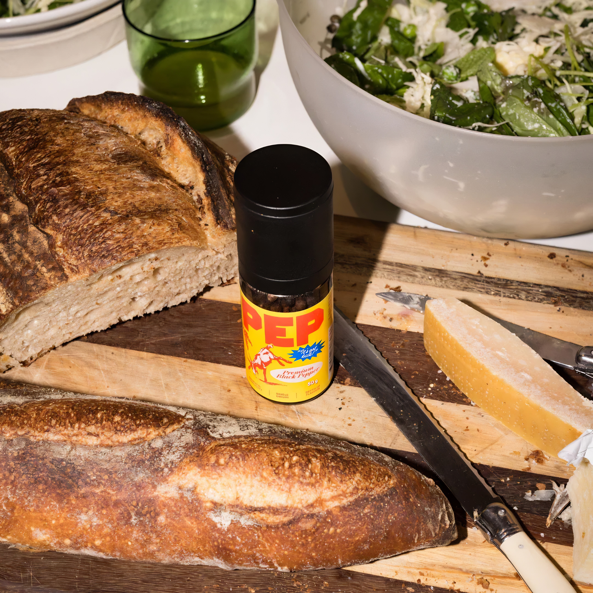 On a chopping board with bread, the PEP Pepper Grinder 50g, for fresh black peppercorns in a reusable smoky glass with yellow cover, with aromatic complexity grown in Cambodia.