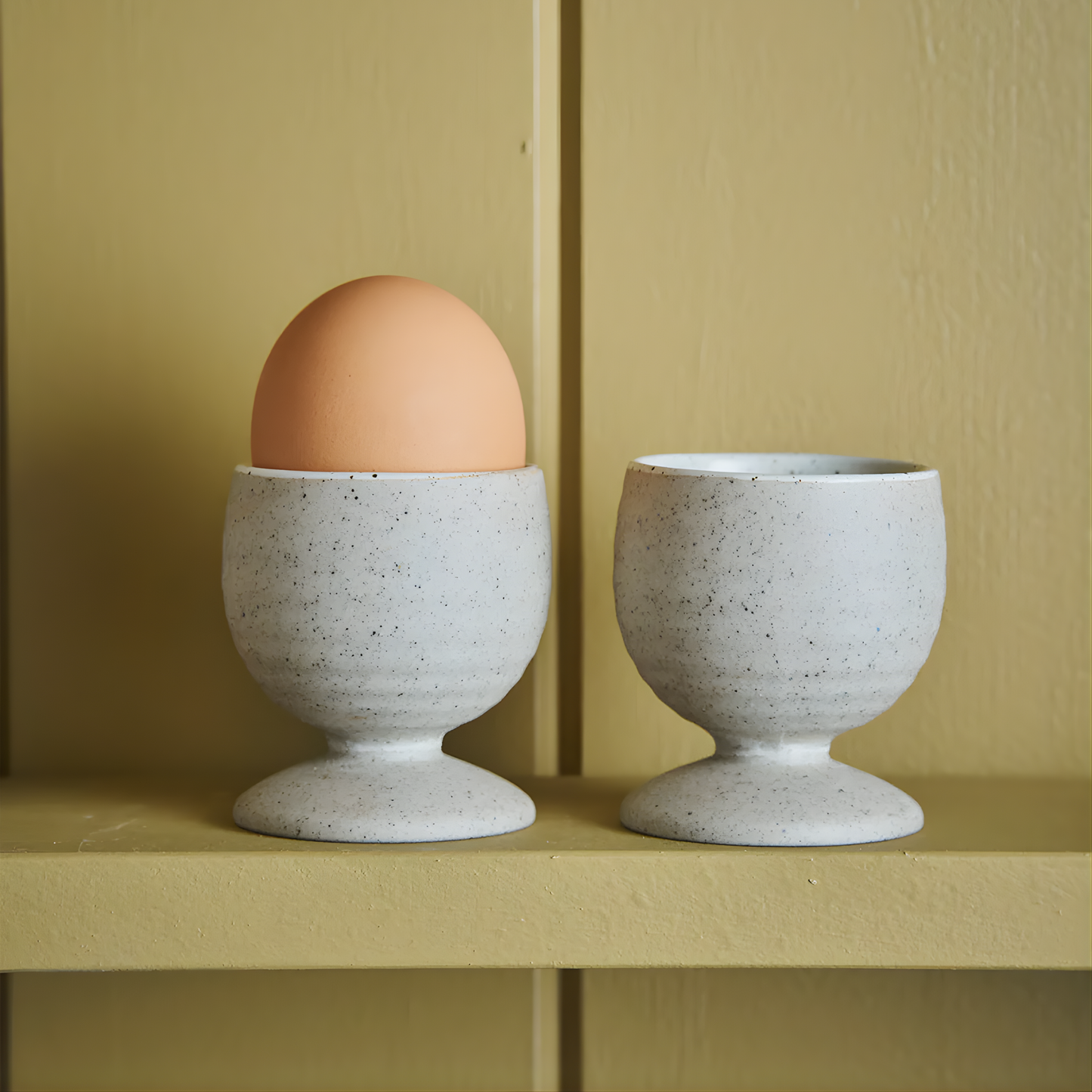 Showing two egg cups on a kitchen cabinet with one having one egg on top. These are the Robert Gordon Garden To Table Egg Cups (set of 2) 5.3cm | 6cm Off-white Earthly clay body with light organic glaze and dark speckles, hand crafted made from stoneware, microwave and dishwasher safe, designed in Australia, made in China.
