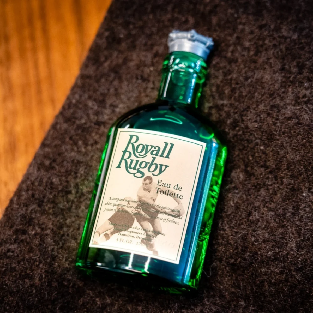 Laid on the table, the Royall Rugby Eau de Toilette by Royall Lyme; green hand-made glass bottle, pewter-colored crown cap, cream label with green text.