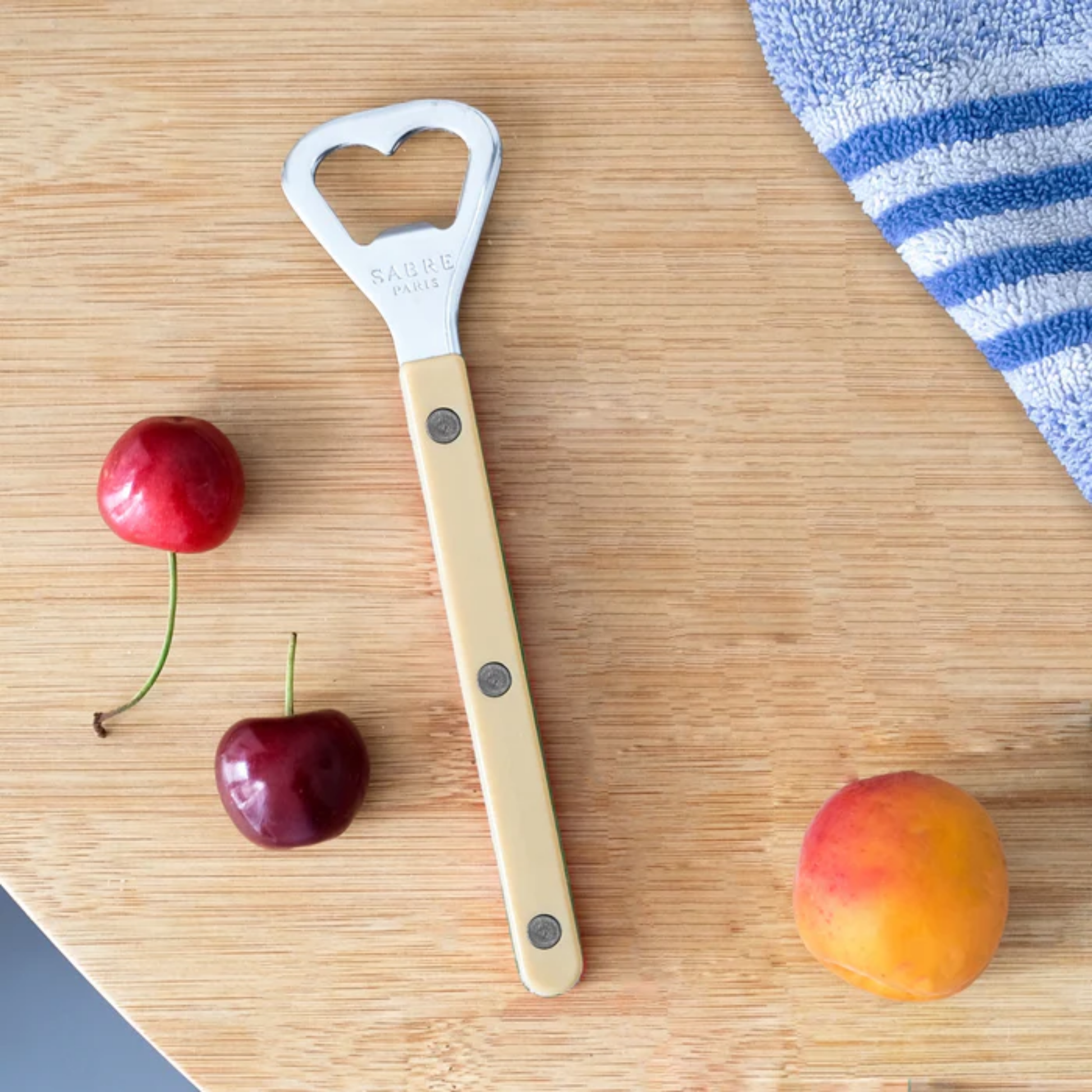 On a table with peach and cherries Sabre Paris Bistrot Bottle Opener 16 cm Ivory - stainless steel and nylon bottle opener with riveted handle, café-inspired design, gloss or vintage finish, perfect for opening bottles with style.