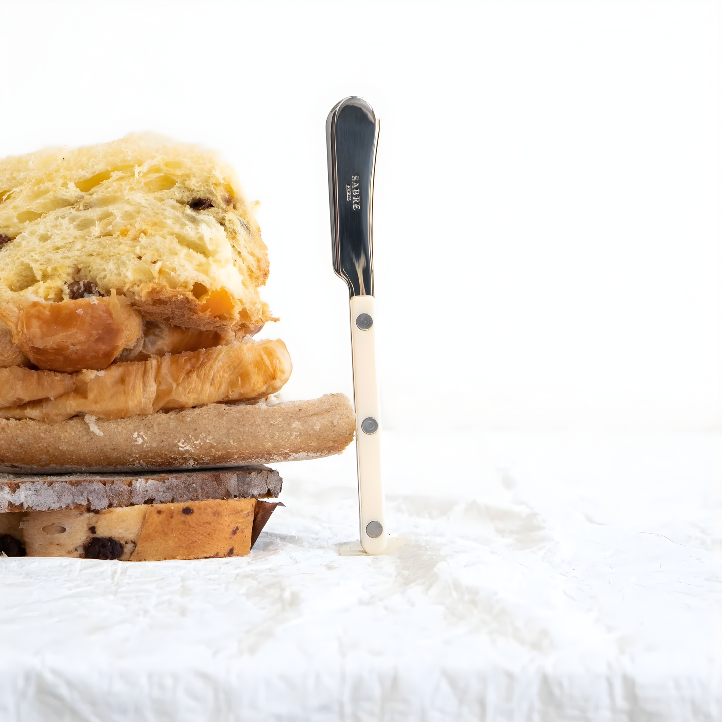 Standing beside a pile of bread, the Sabre Bistrot Cutlery Butter Spreader Ivory 14cm, which is made from Inox 18/10 Manche, features a non-slicing blade, perfect for spreading soft butter, tapenades, taramas, and other spreads on bread or toast, dishwasher safe at 45°C maximum and not suitable for microwave oven.