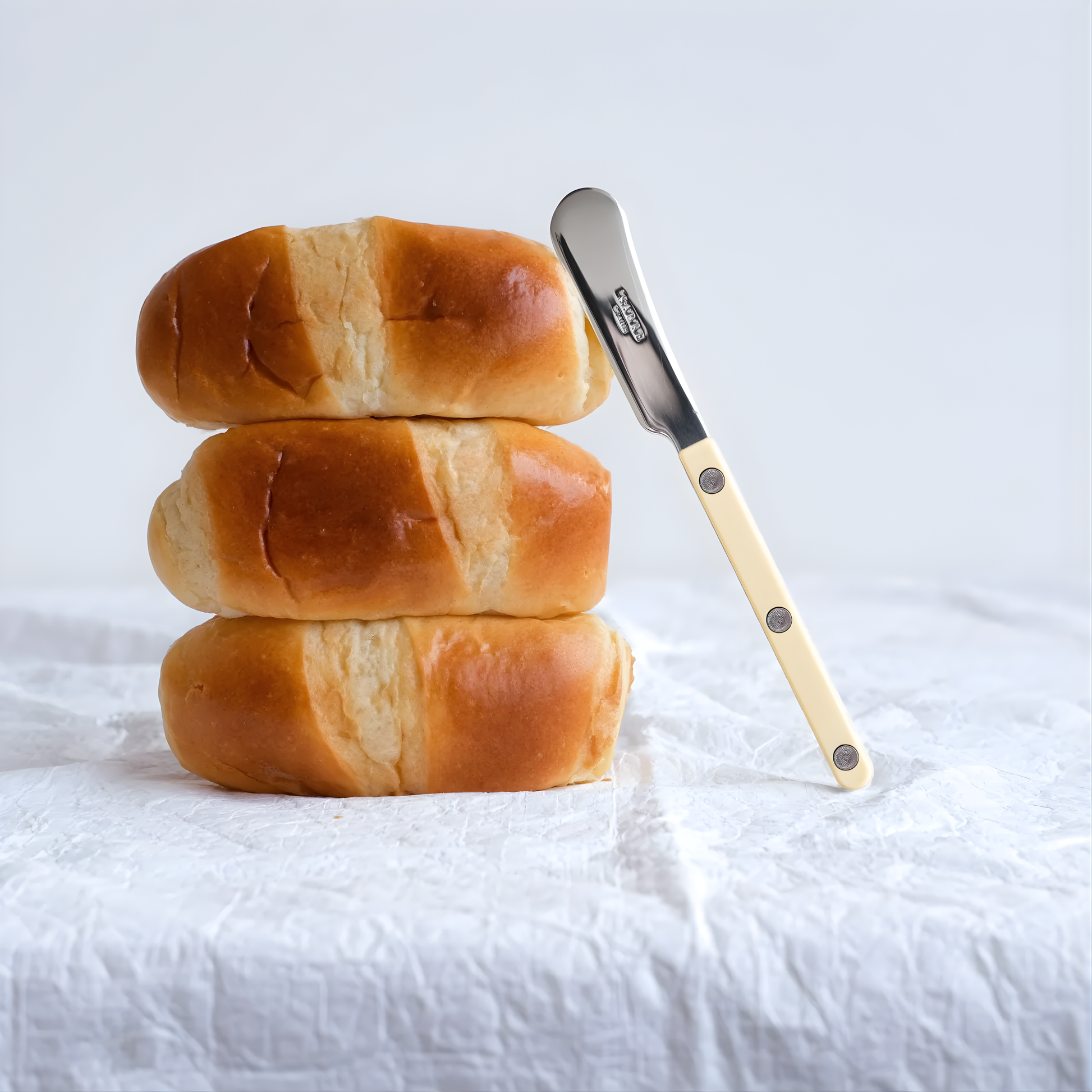 Leaning on a pile of bread, the Sabre Bistrot Cutlery Butter Spreader Ivory 14cm, which is made from Inox 18/10 Manche, features a non-slicing blade, perfect for spreading soft butter, tapenades, taramas, and other spreads on bread or toast, dishwasher safe at 45°C maximum and not suitable for microwave oven.